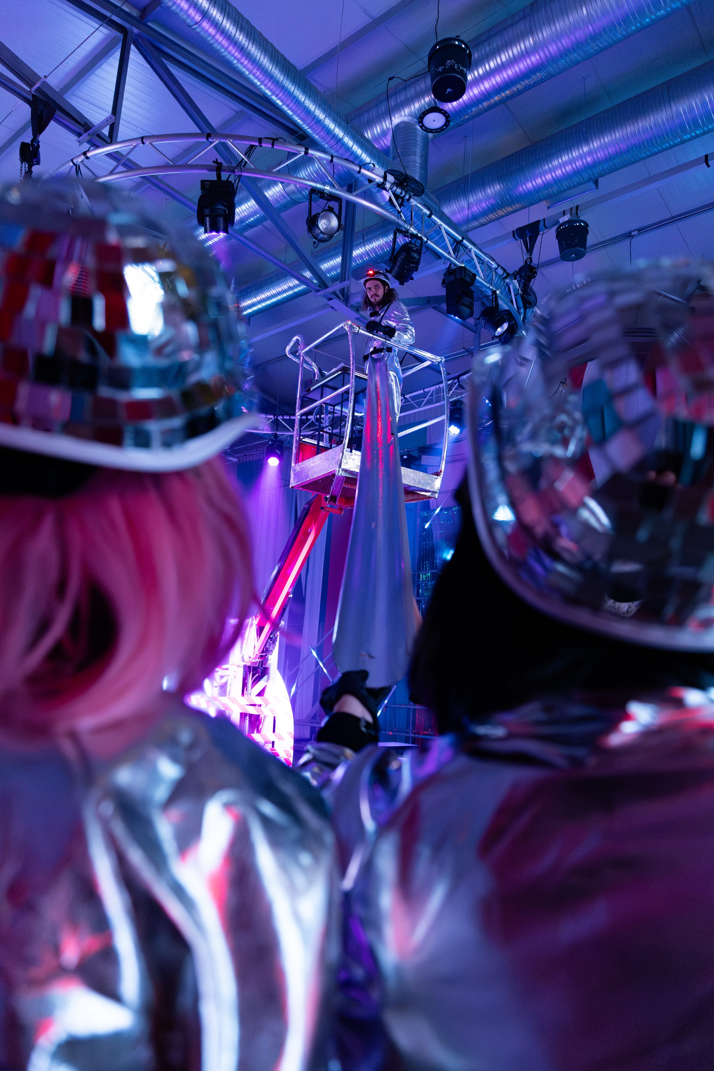 Person dressed as a space explorer standing on a raised platform in a room with metallic ductwork and stage lighting, surrounded by reflective disco balls.
