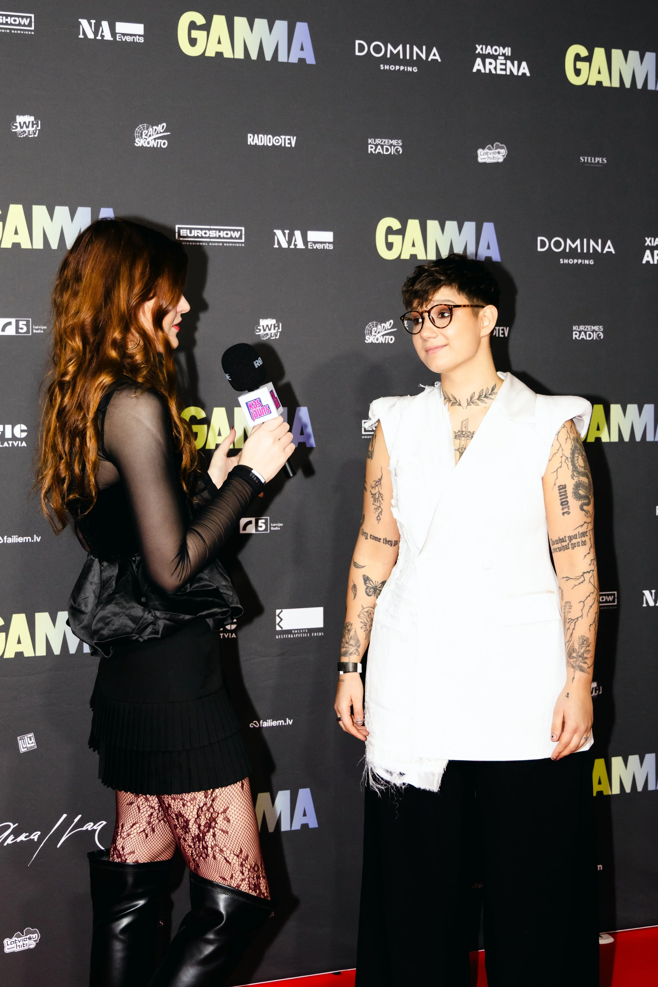 A woman in black attire and fishnet stockings interviews a woman with short dark hair, glasses, and tattoos at a media event with a black backdrop featuring various logos and the word GAMMA.