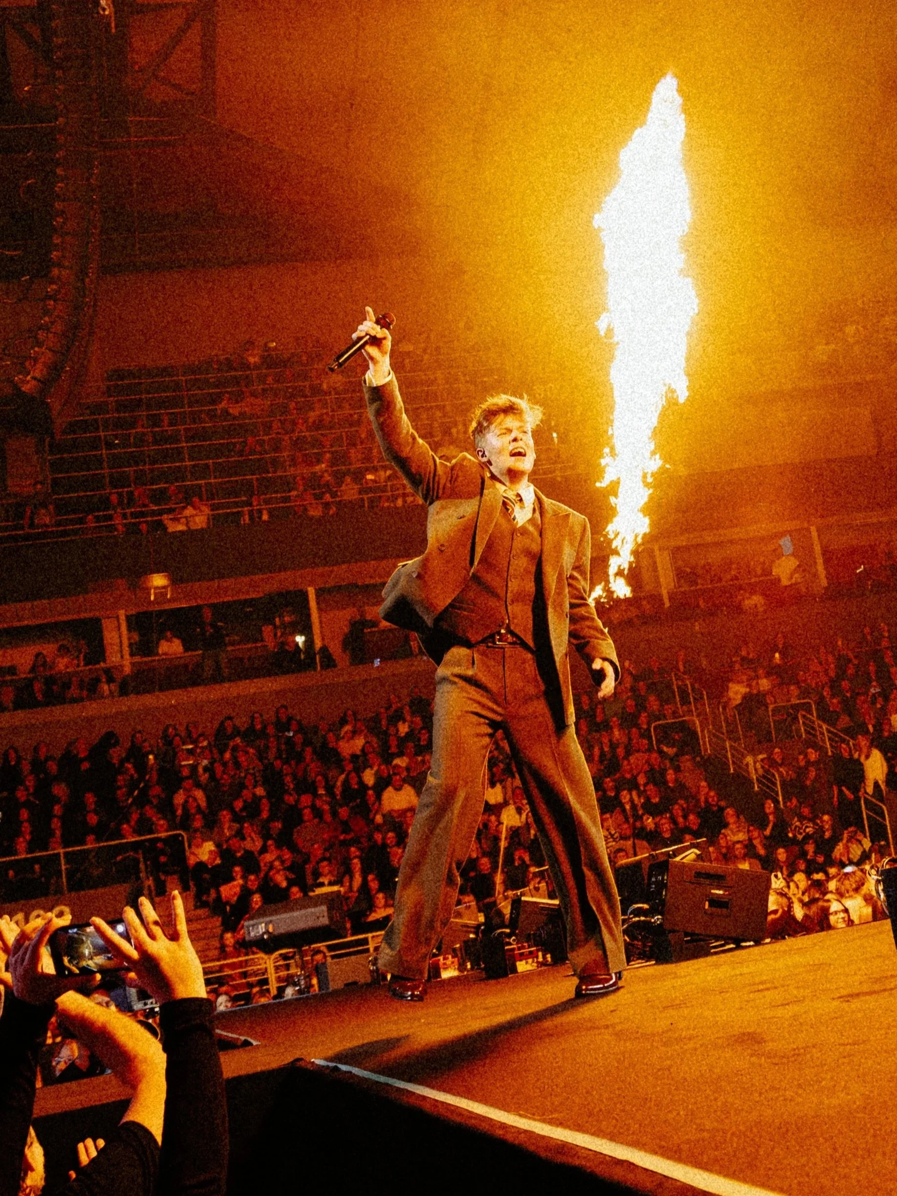 A performer in a jacket and pants holding a microphone on stage in front of a crowd, with fire shooting up from the stage.