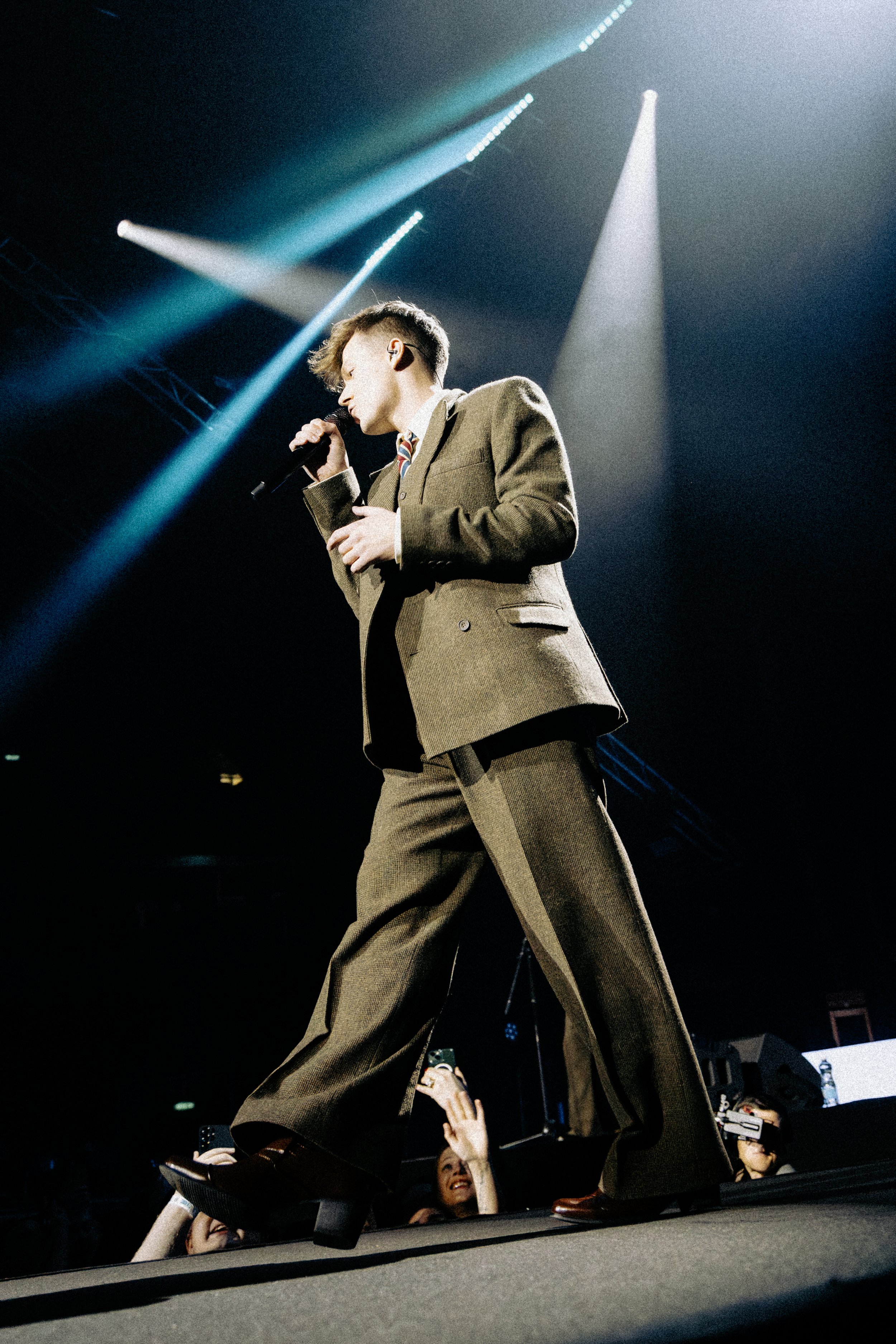 Male performer with a microphone on stage under bright lights, wearing a brown suit.