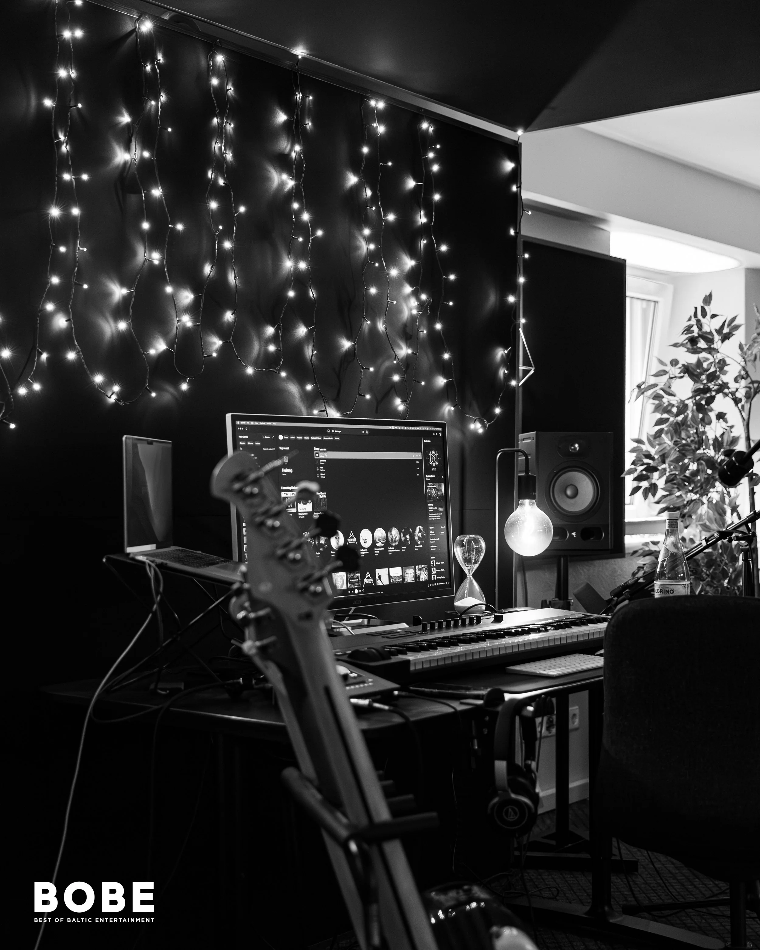 A music or recording studio setup with a computer monitor displaying a music library, a MIDI keyboard, a pair of studio monitors, and decorative string lights on the black wall.