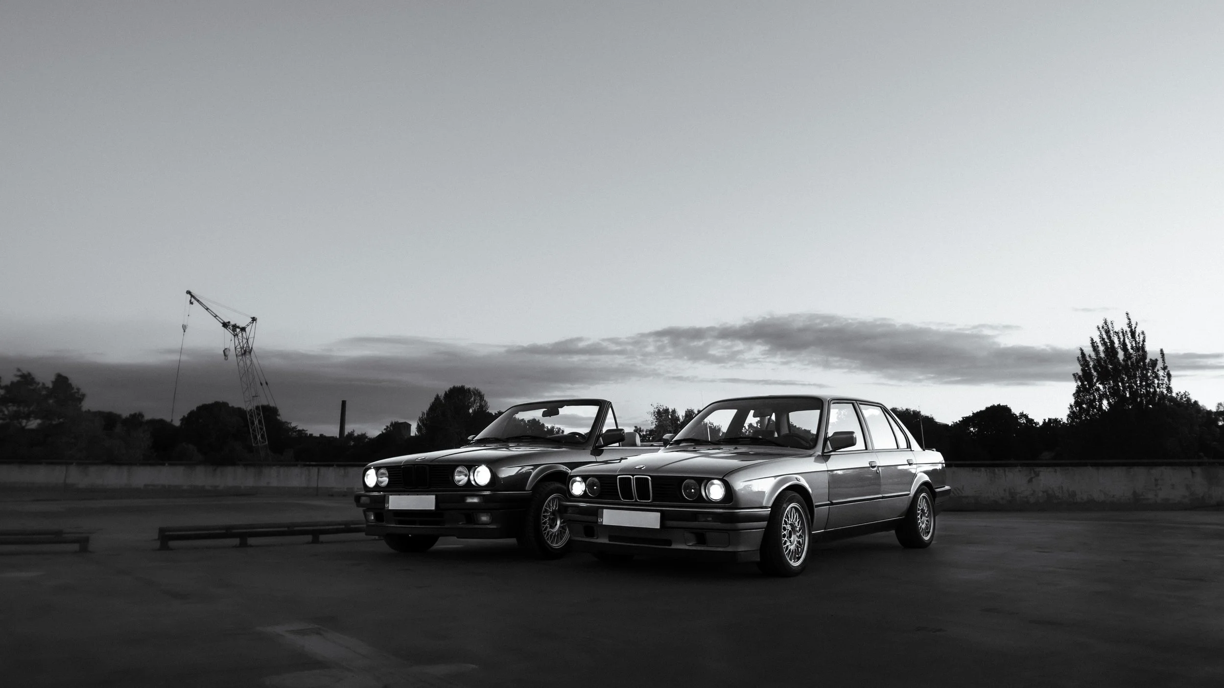 Black and white photo of two vintage BMW cars parked next to each other on a rooftop parking lot with a city skyline and construction crane in the background.