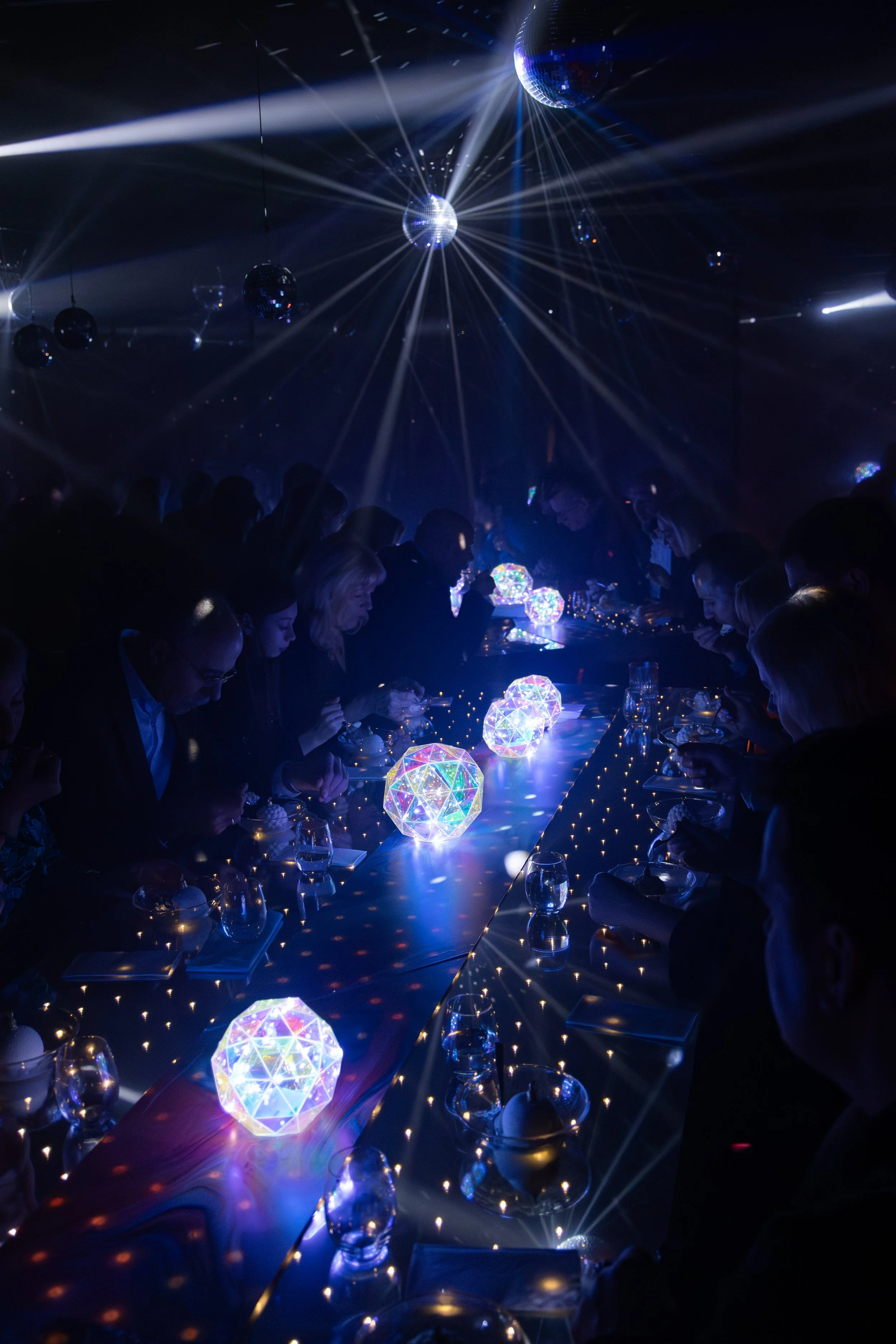 People are gathered in a dark room with a long table decorated with glowing, multi-faceted illuminated geometric shapes. Multiple disco balls hang from the ceiling, reflecting light beams across the scene, creating a lively and festive atmosphere.