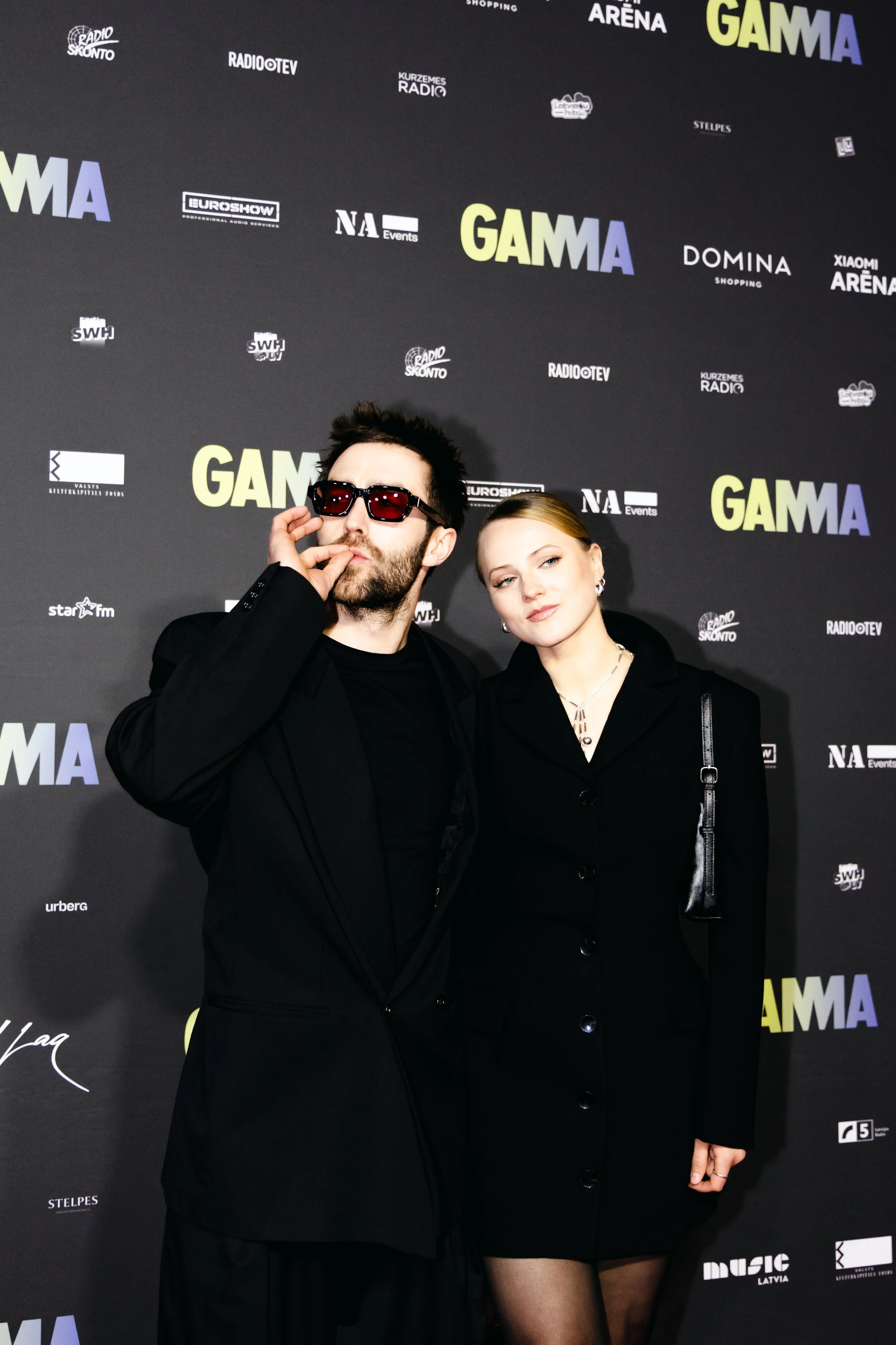 Two people, a man and a woman, posing together at a dark-themed event. The man is wearing sunglasses and a black suit, touching his face with one hand. The woman is dressed in a black blazer and has blonde hair, holding a handbag. They are standing i