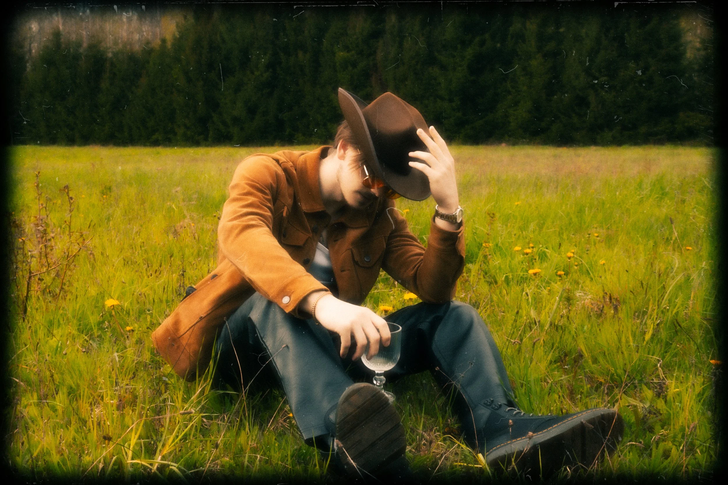 A person sitting in a grassy field holding a plastic cup, wearing a brown jacket, black hat, and sunglasses, with trees in the background.