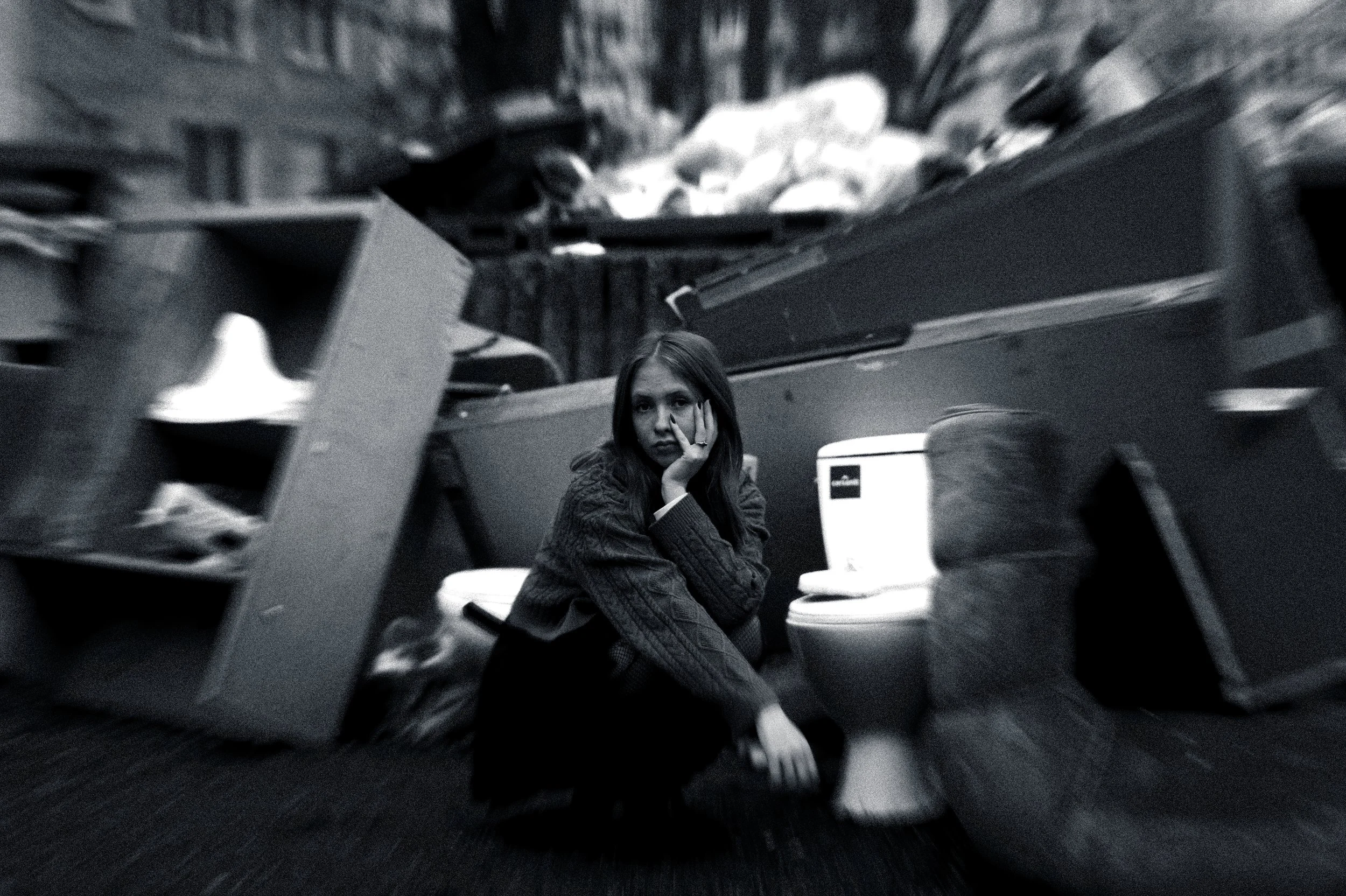 A young woman sitting in a room, resting her face on her hand, looking contemplative or bored, with various objects around her and a blurry background.