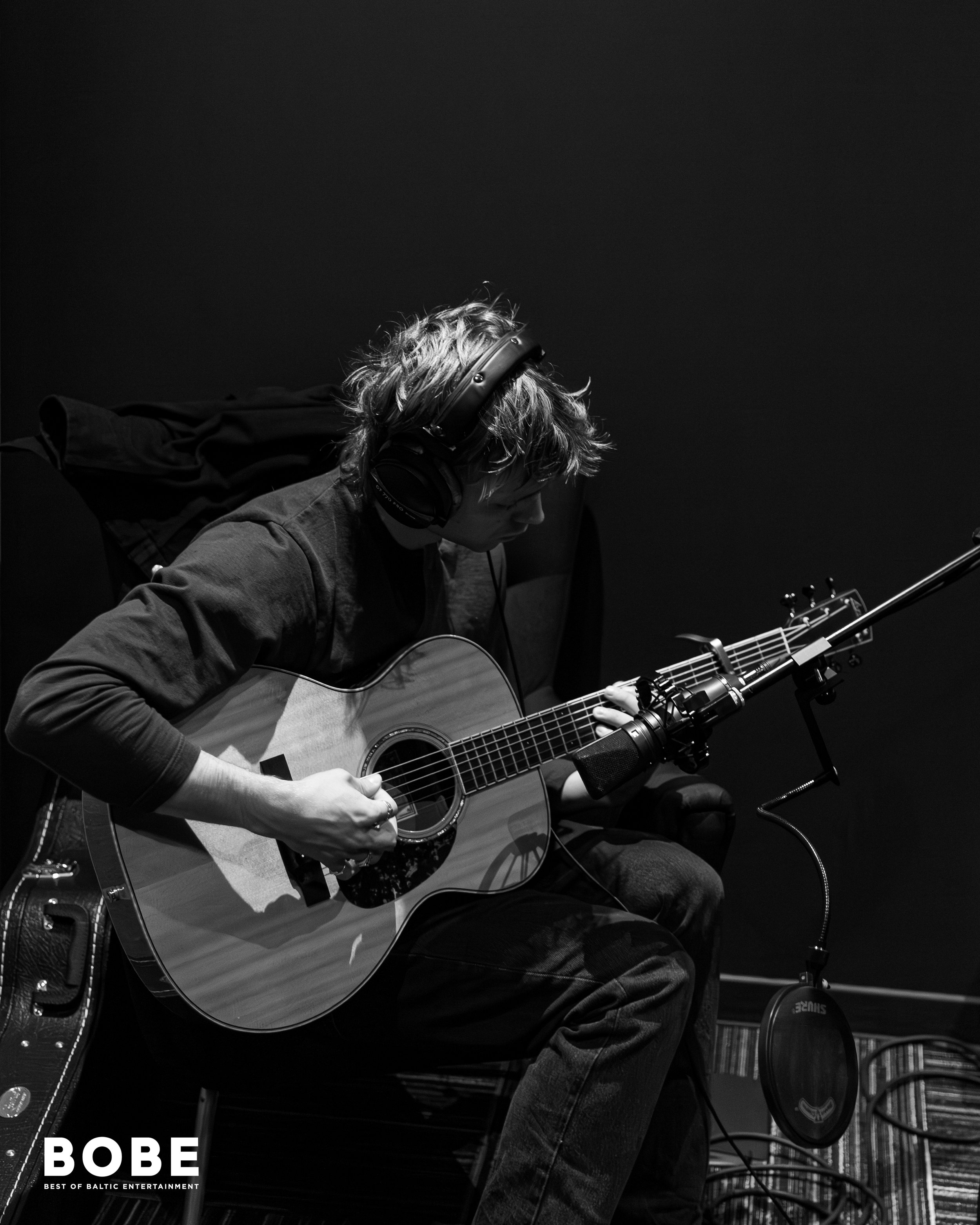A person with wavy hair, wearing headphones, playing an acoustic guitar, photographed in black and white in a music studio or recording space.