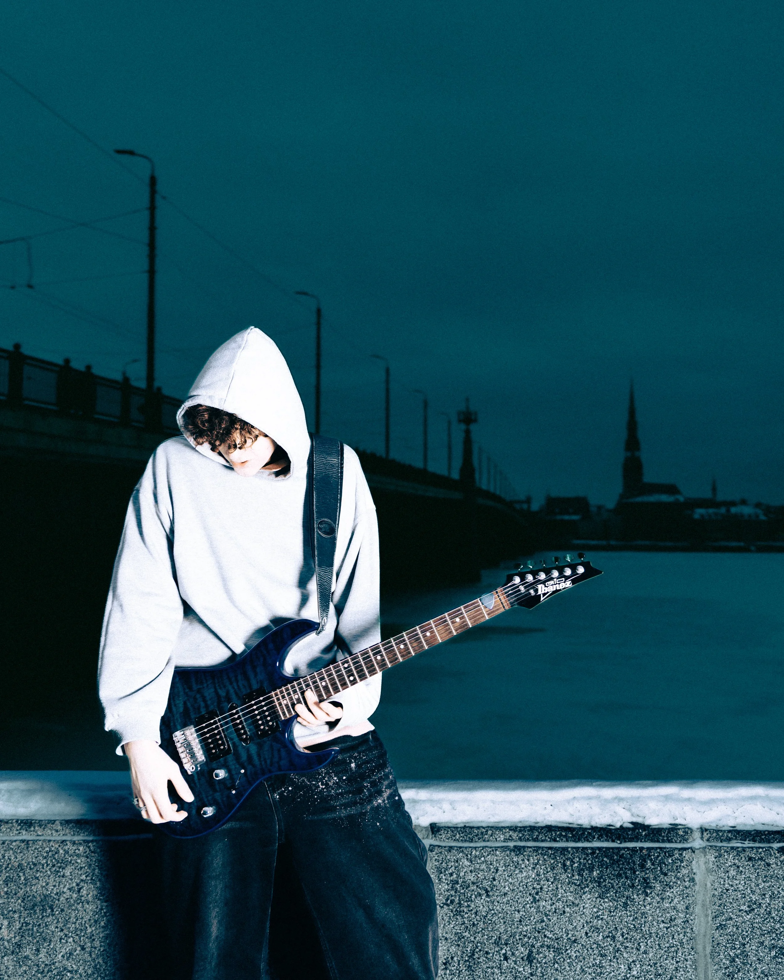A young person wearing a light gray hoodie and dark jeans, playing a blue electric guitar outdoors during dusk, with a river, bridge, streetlights, and silhouette of a church steeple in the background.