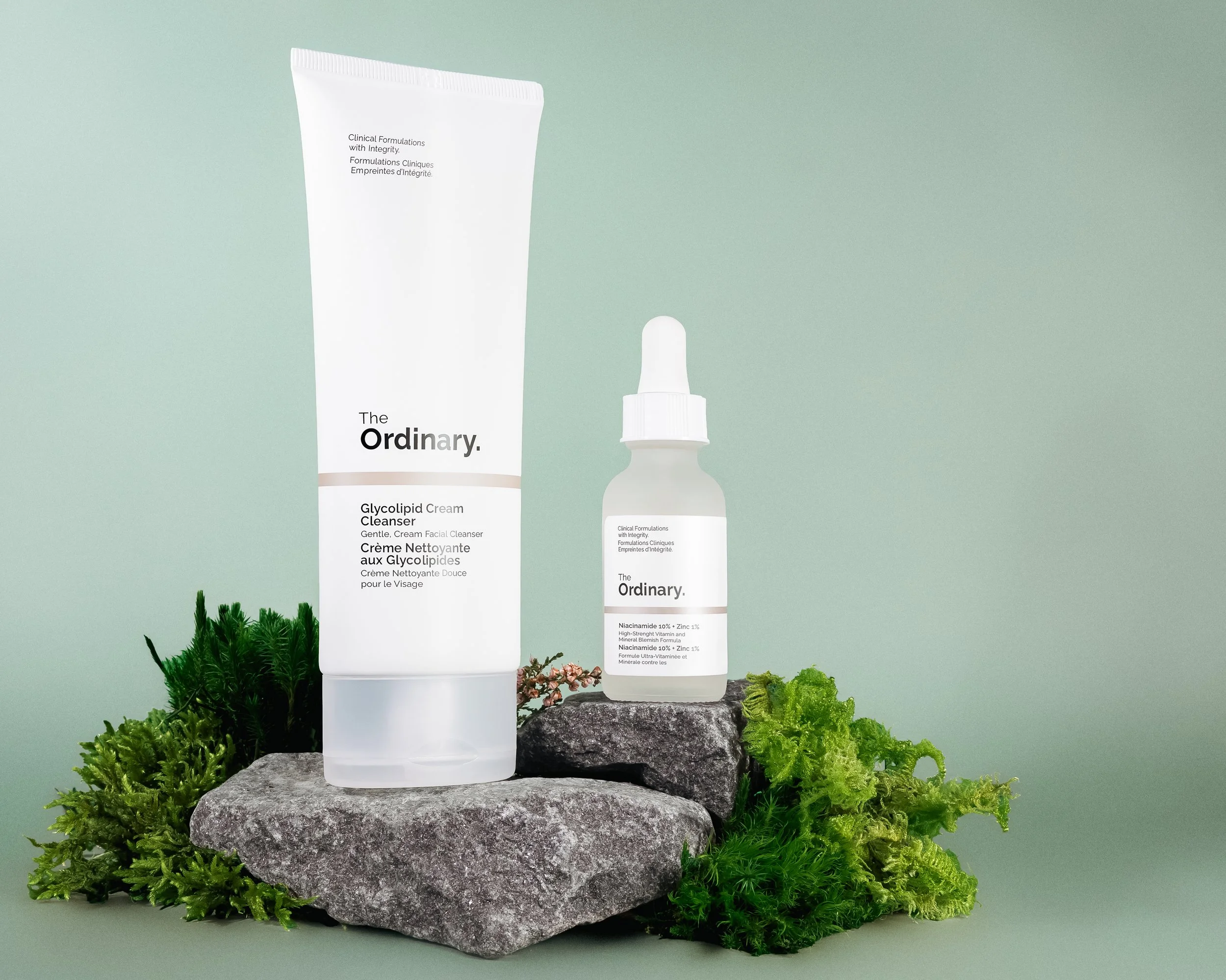 Two skincare products from The Ordinary brand, set on a rock with green plants around them, against a plain light green background.