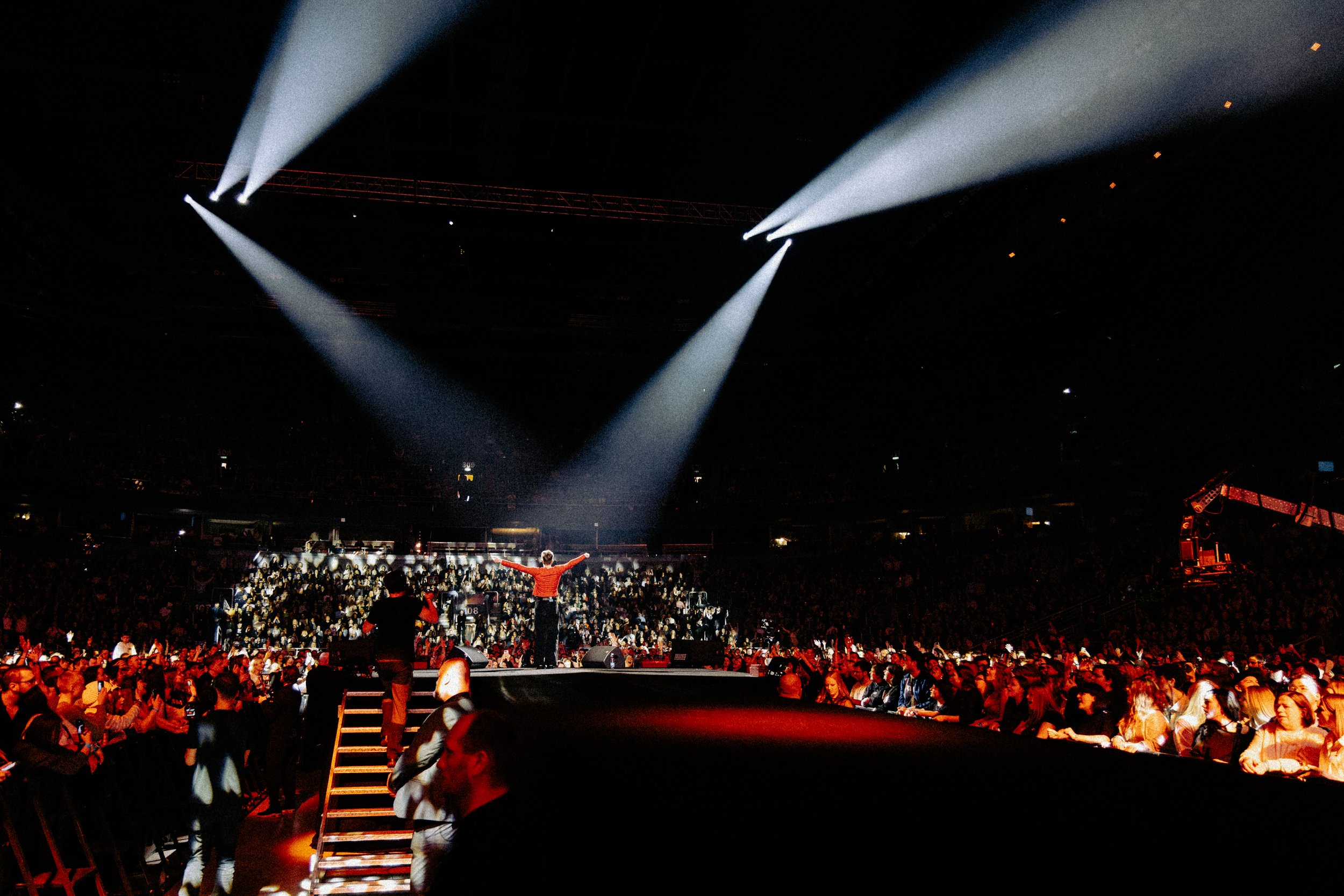 A performer with arms outstretched on stage in front of a large audience at a concert or event. Bright spotlights shine down from above, creating a dramatic lighting effect amidst a dark background.