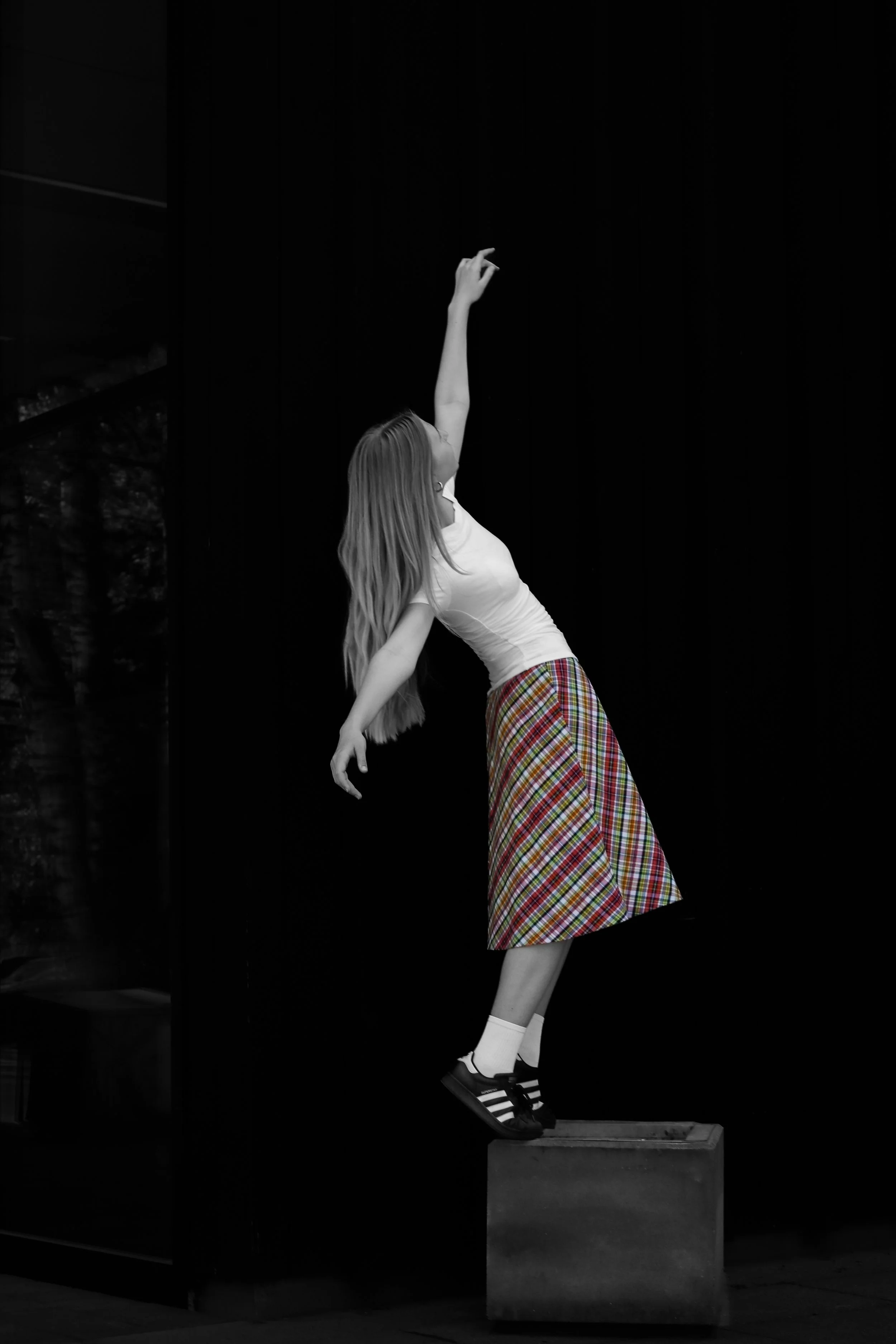 A woman with long hair wearing a white top and plaid skirt is balancing on a wooden platform, with one arm extended upward and the other downward, against a dark background.