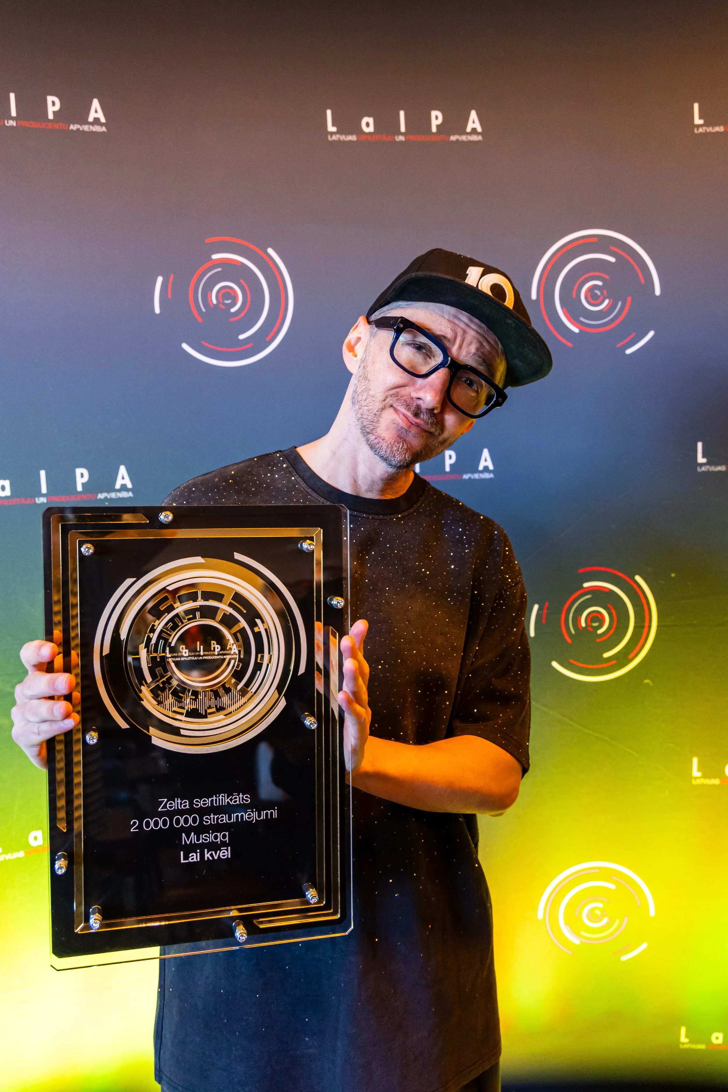 A man with glasses and a black cap is holding a large framed certificate in front of a colorful background with circular designs. The certificate has Latvian text and mentions a donation of 2,000,000 lats to a music school.