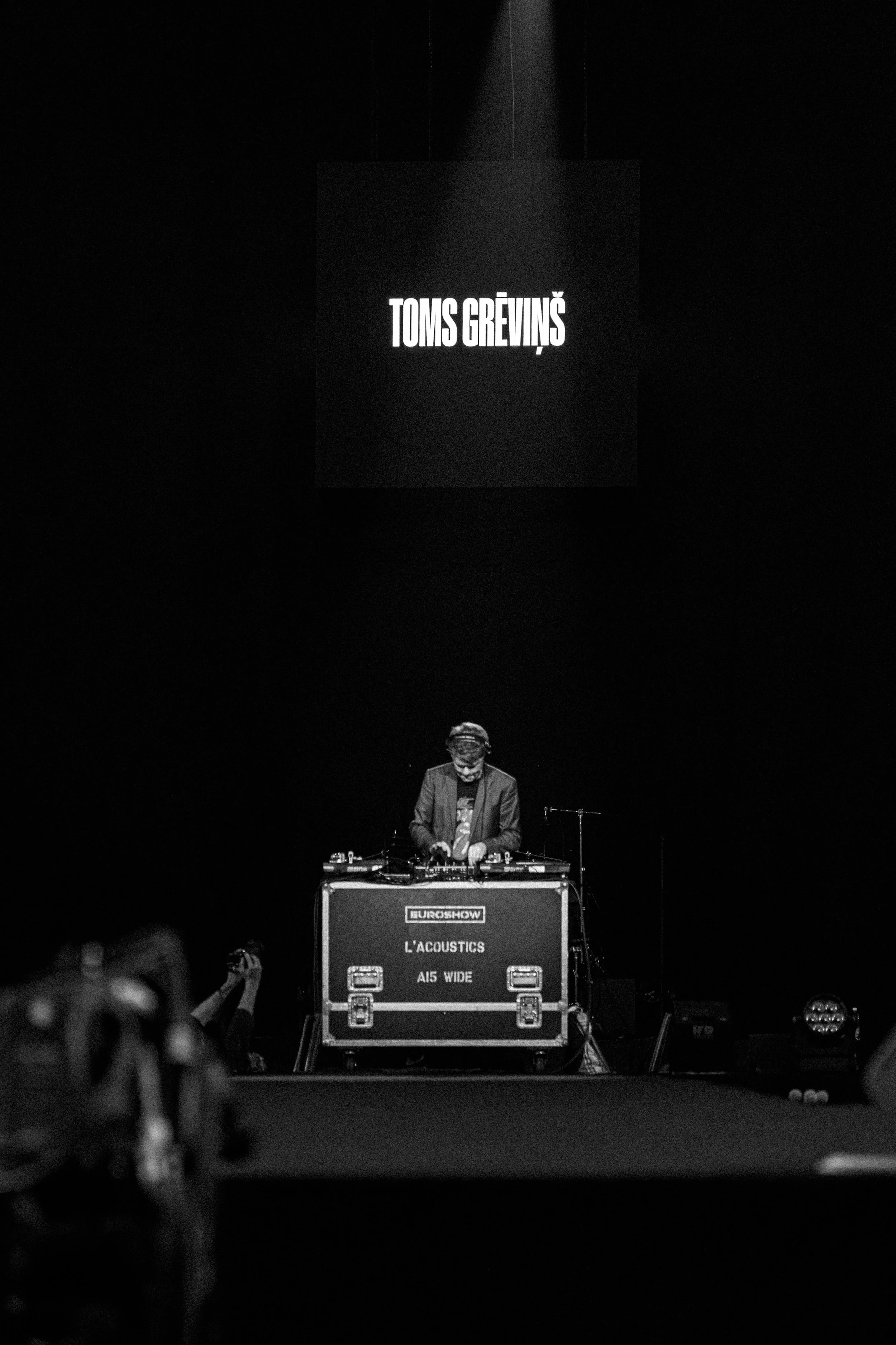 A DJ performs on stage with a large black screen behind him displaying 'TOMS GREVINŠ'. The DJ is focused, standing behind a setup that includes a table with DJ equipment, and is surrounded by a dark audience setting.