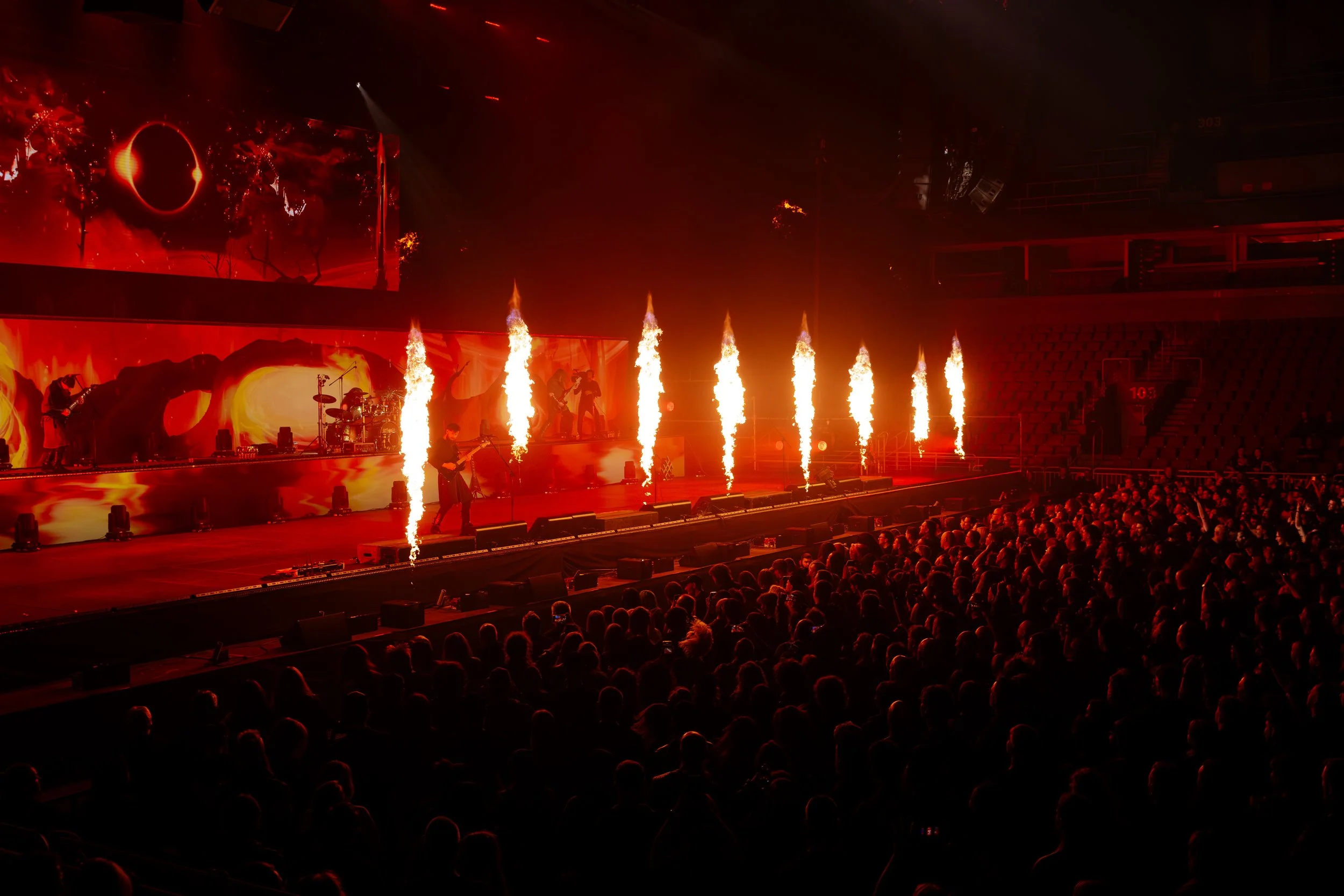 A live concert stage with musicians performing, pyrotechnics shooting upwards, and a large LED screen displaying an red and black abstract image, all illuminated in red lighting, with an audience watching from the darkened seats.