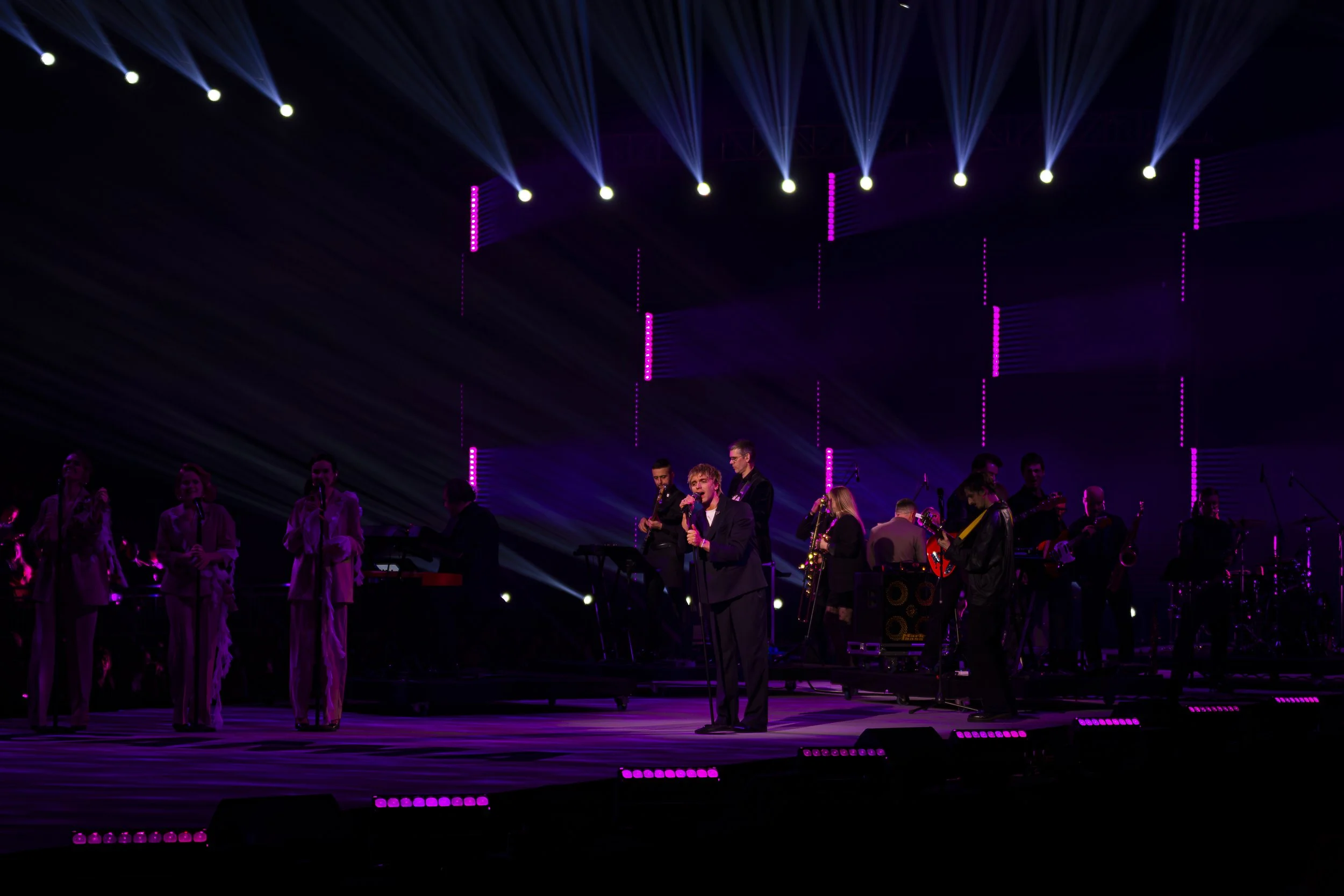 A musical performance on stage with multiple singers and musicians under purple and blue stage lights.