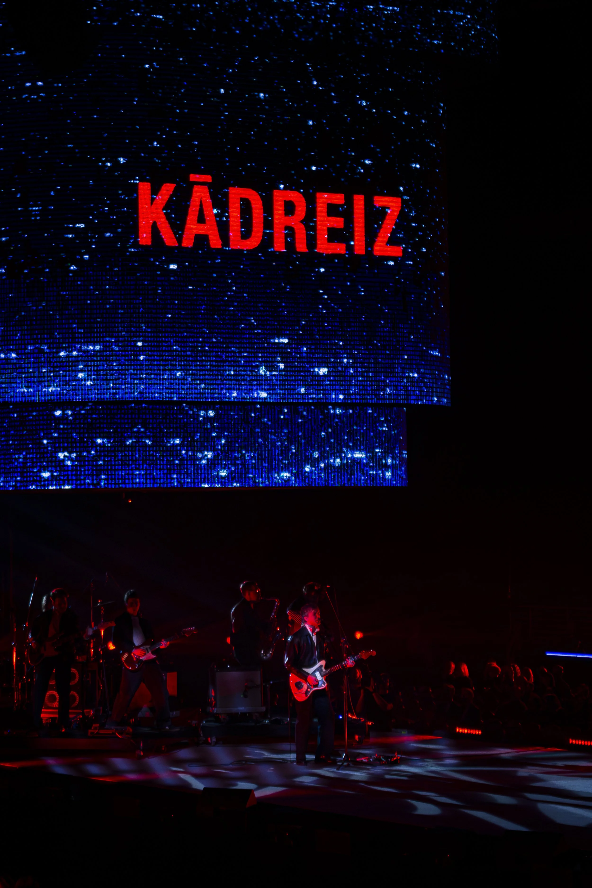 Musicians perform on stage at night with a large digital screen showing the word 'KADREIZ' in red letters in the background.