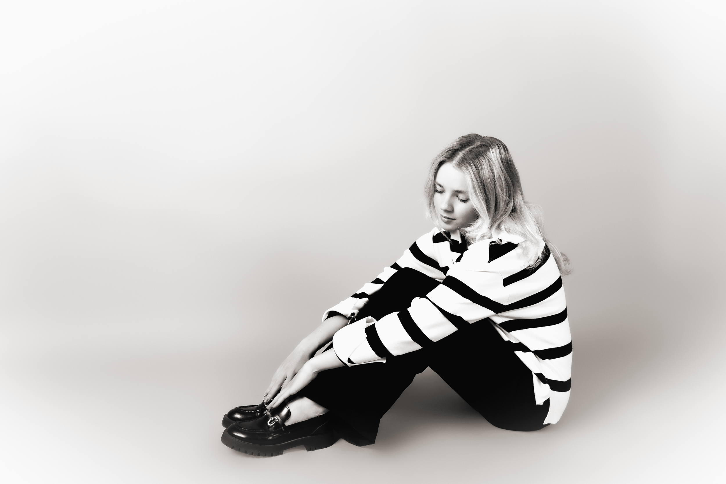 Black-and-white photo of a young woman with long wavy hair sitting on the floor, wearing a striped long-sleeve shirt and shiny black boots, against a plain light-colored background.