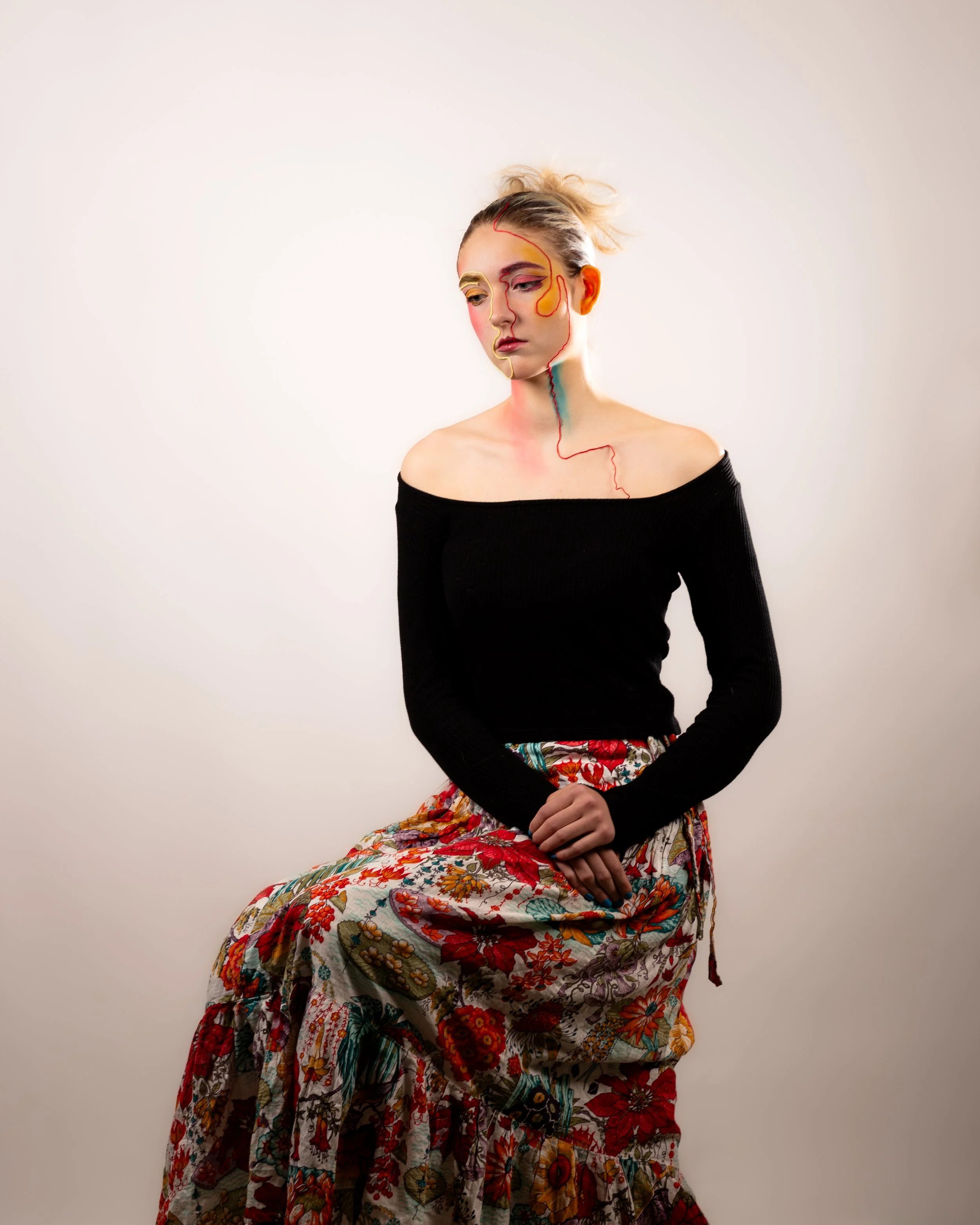 A woman with a pensive expression, wearing a black off-the-shoulder top and a colorful floral skirt, sitting against a plain white background, with artistic line drawings on her face in various colors.