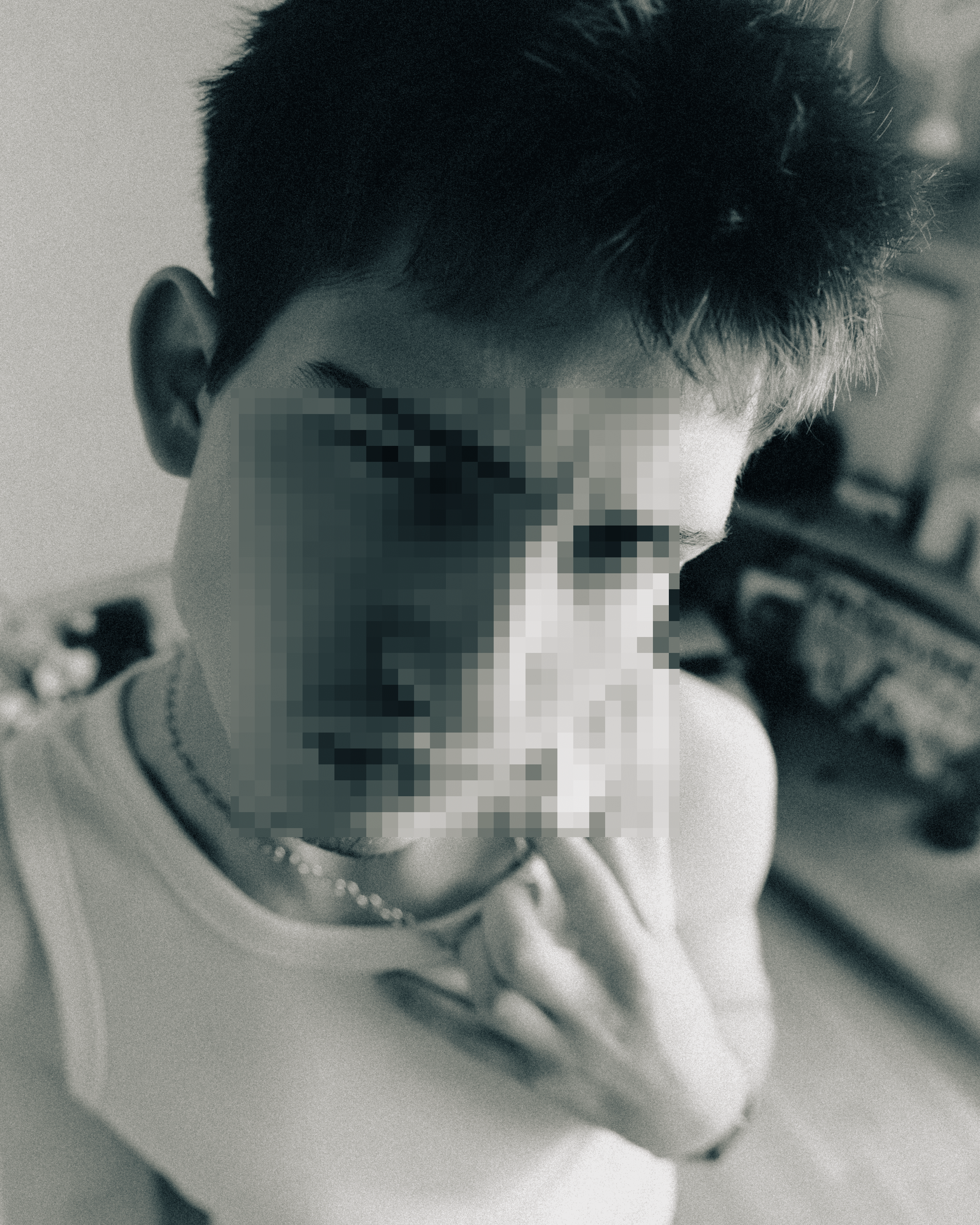 Close-up of a person's face with pixelation over the eyes, showing dark short hair, wearing a necklace, with a blurred background.