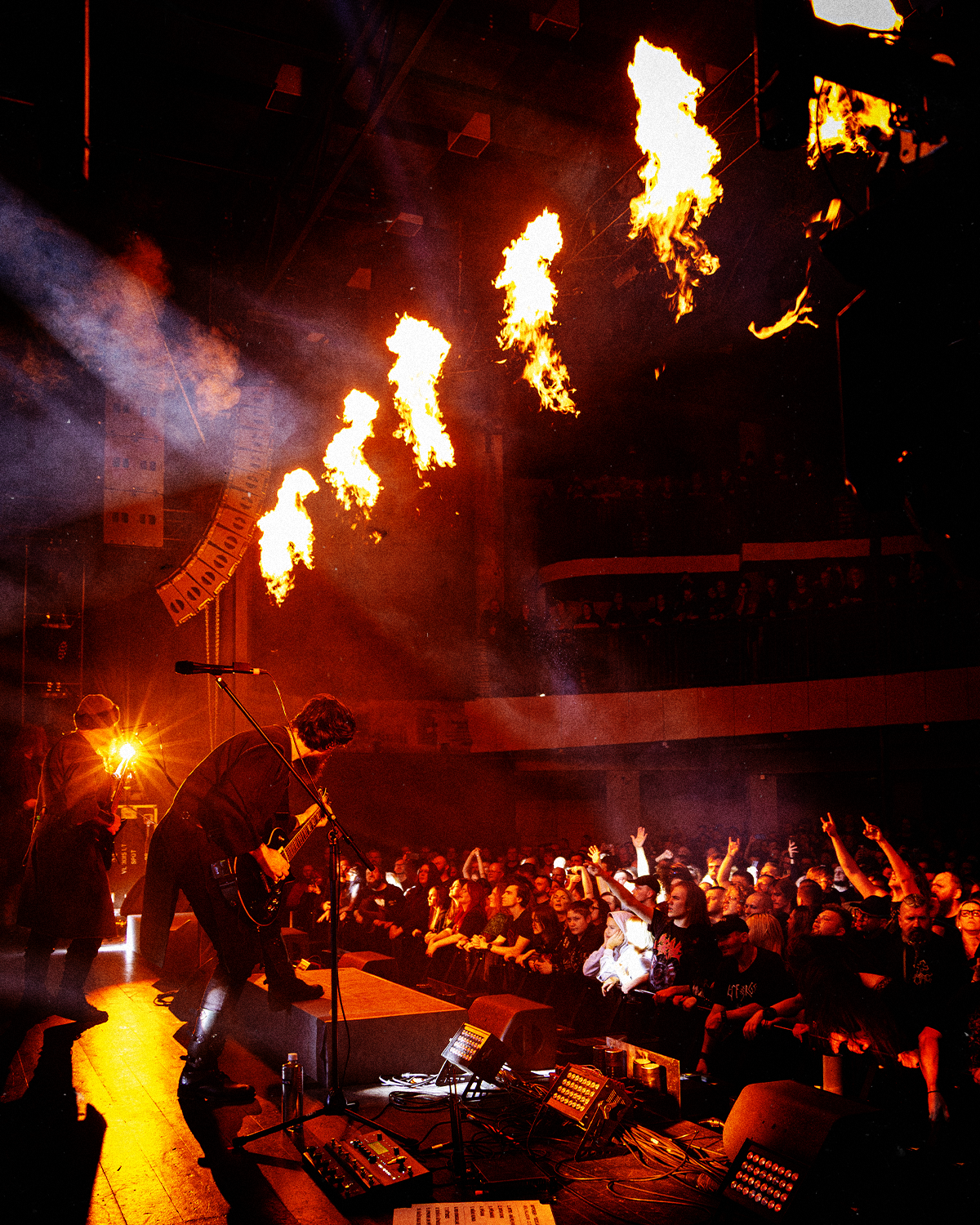 A live concert with a band performing on stage, flames shooting from overhead pyrotechnics, and an audience enjoying the show in a dark indoor venue.