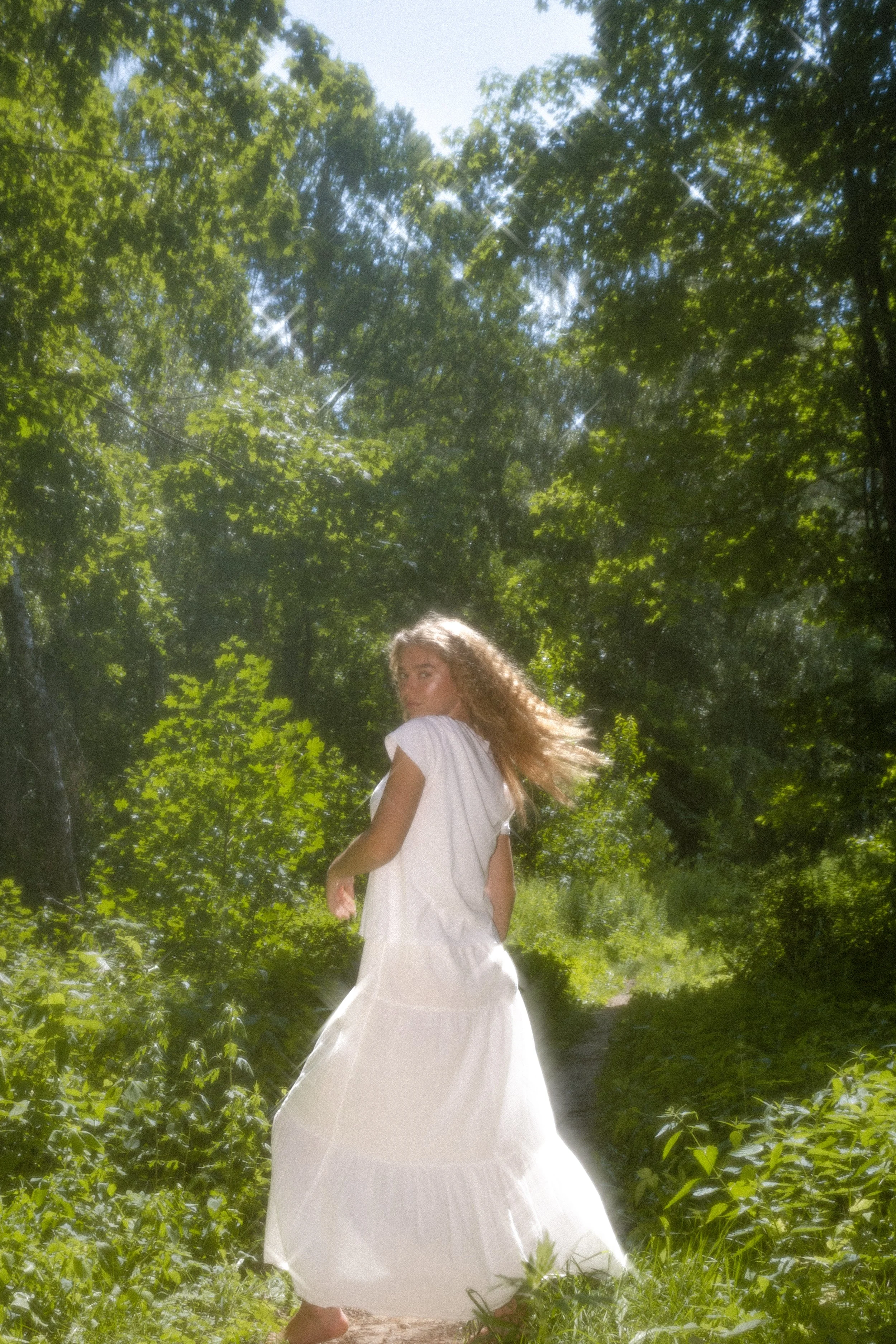 Young woman with long curly hair walking in a lush green forest, wearing a white dress.
