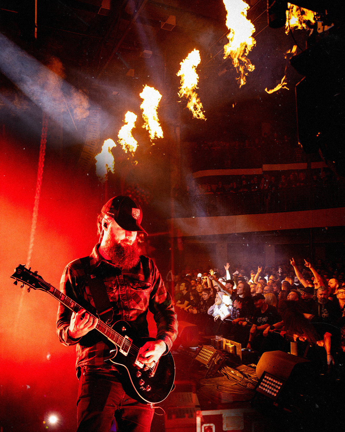 A musician with a beard wearing a baseball cap plays an electric guitar on stage in front of a lively audience, with flames shooting from above, illuminated by red and orange stage lights.