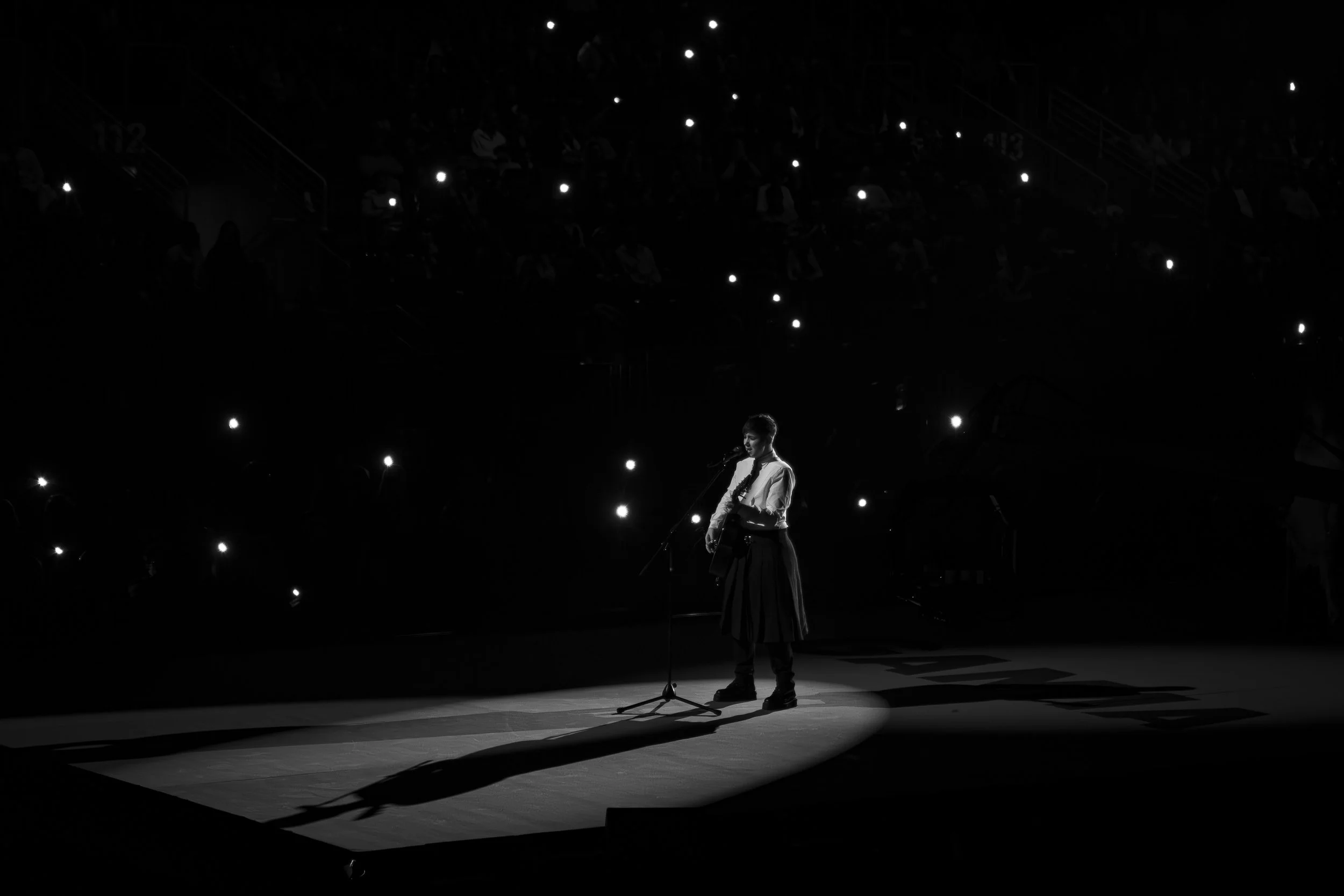 A person stands on stage, playing a guitar and singing into a microphone, illuminated by a spotlight, with a dark background dotted with small bright lights.
