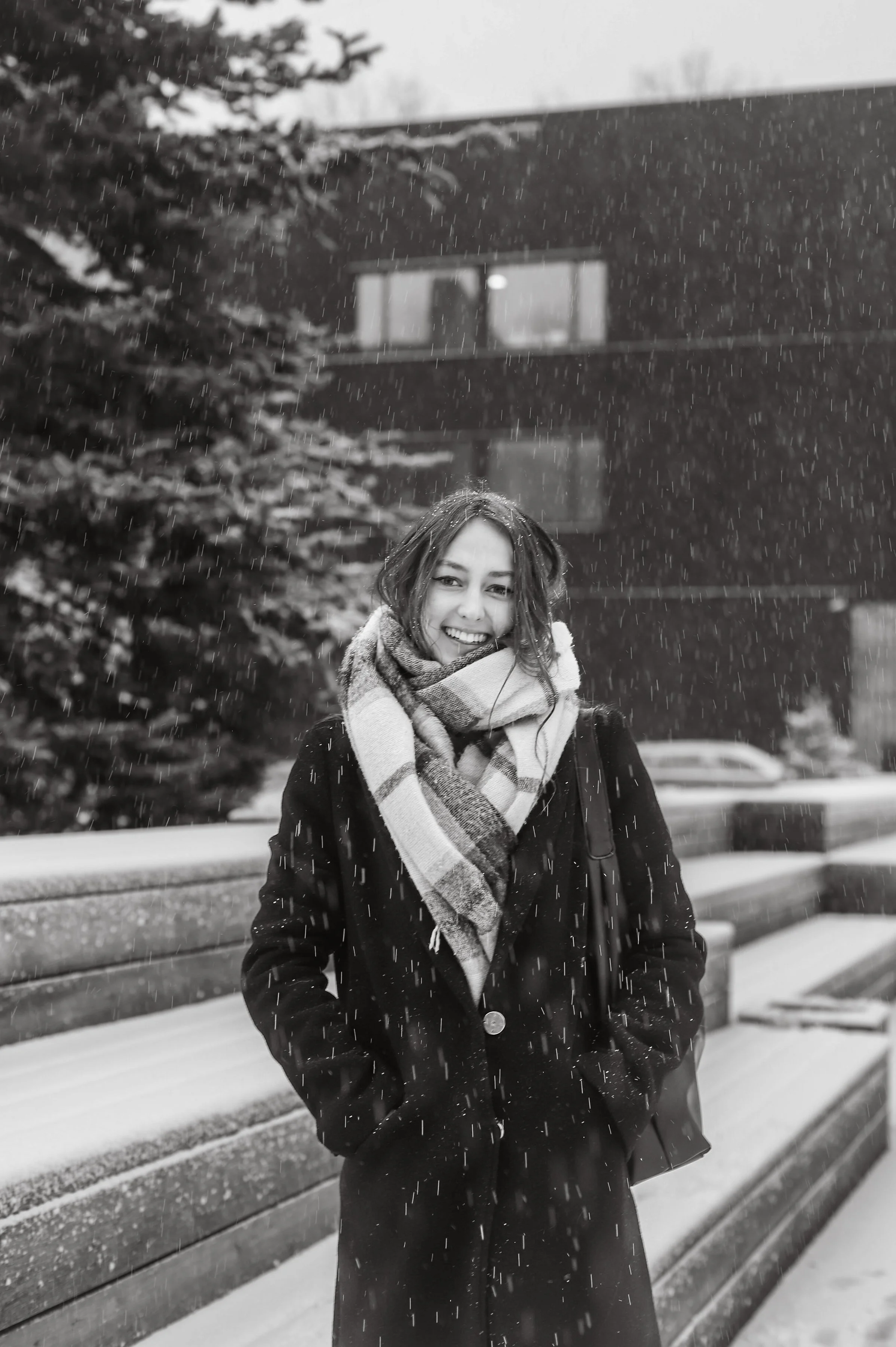 A smiling woman with long hair, wearing a coat and a plaid scarf, stands outdoors in the snow, with snow falling around her, and an apartment building and snow-covered trees in the background.