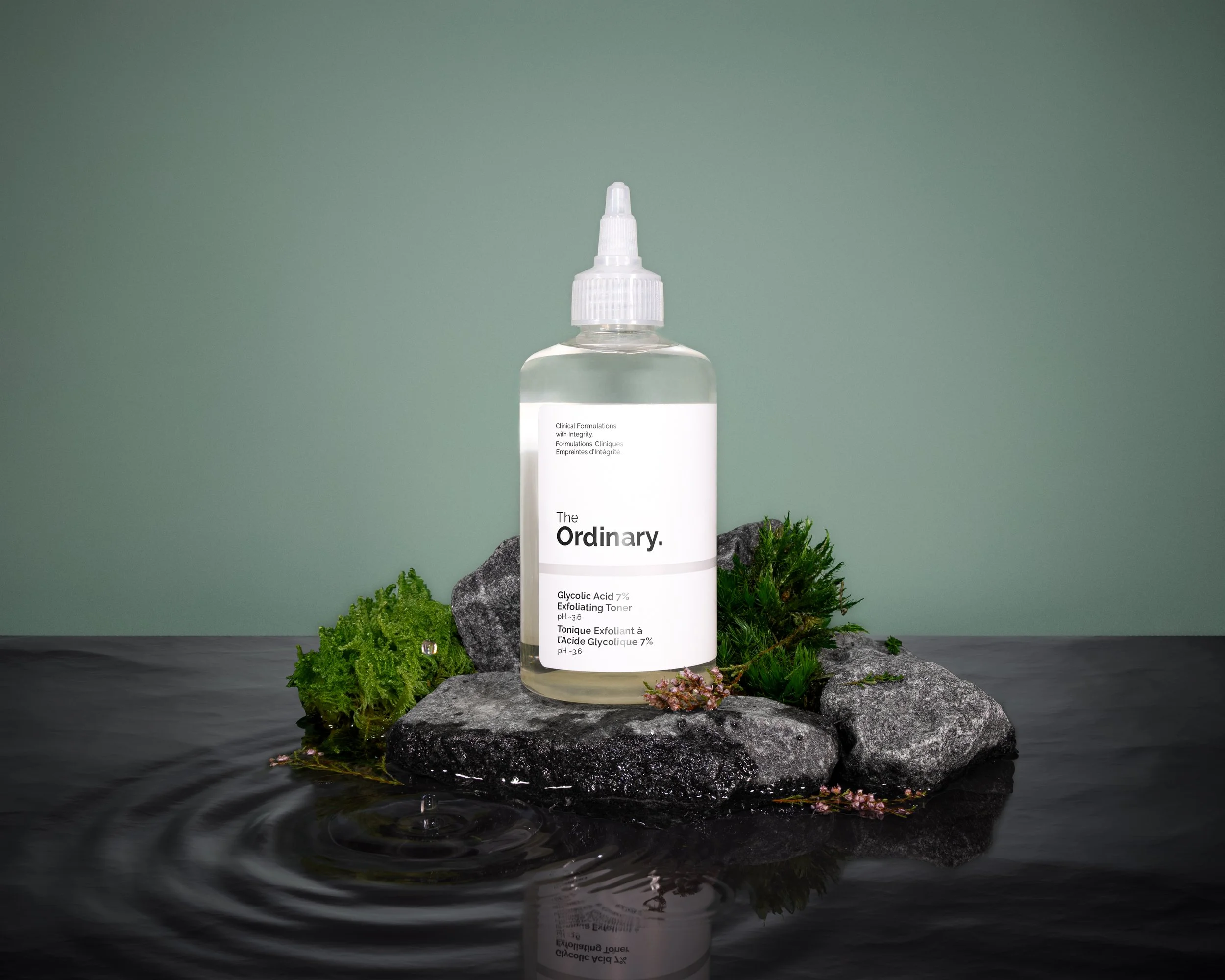 A clear plastic bottle of skincare toner with a white label, placed on rocks surrounded by green plants and flowering moss, with a calm water surface in the foreground and a plain green background.