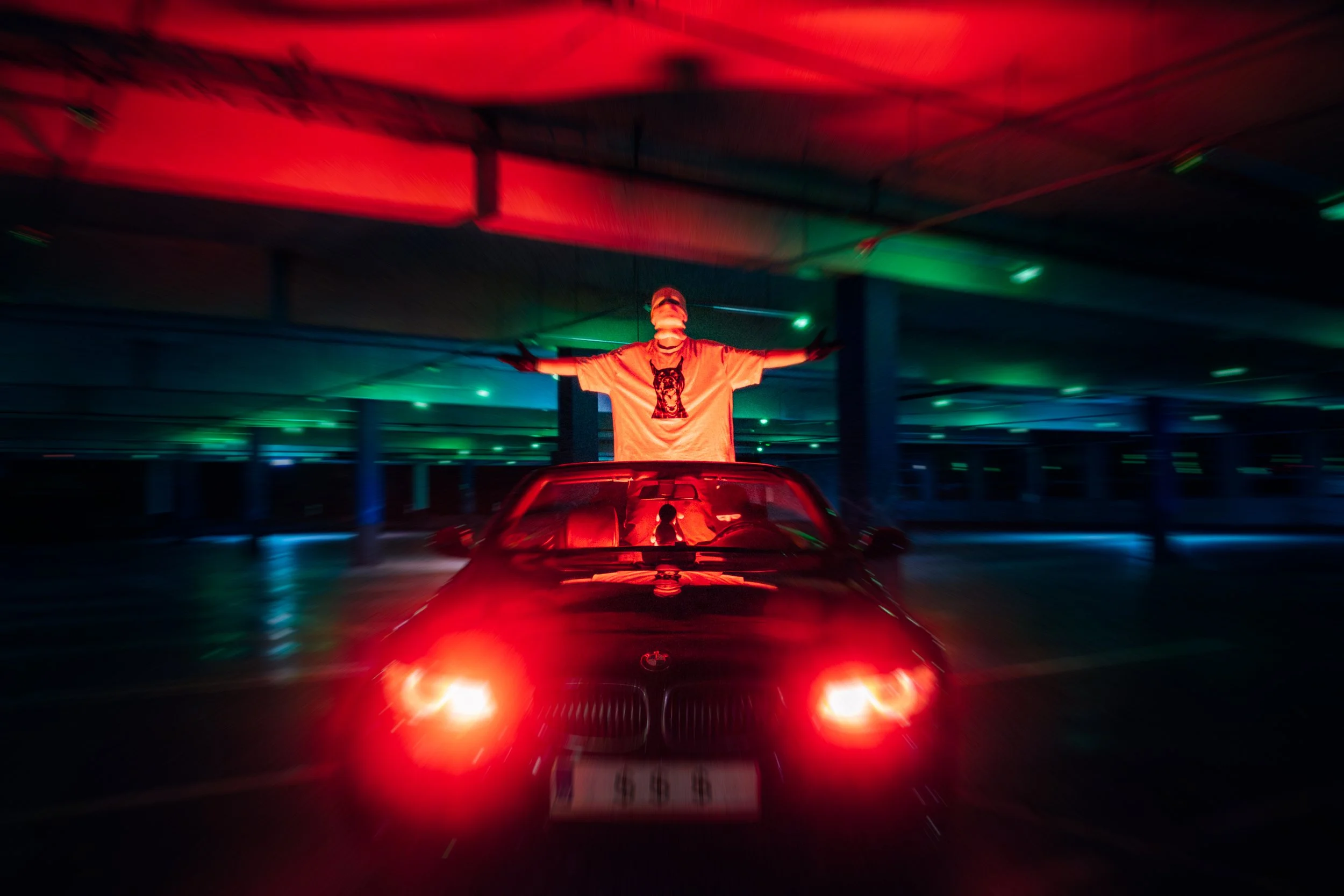 Person standing with arms outstretched on the roof of a red car in a dimly lit parking garage illuminated by red and green lights.