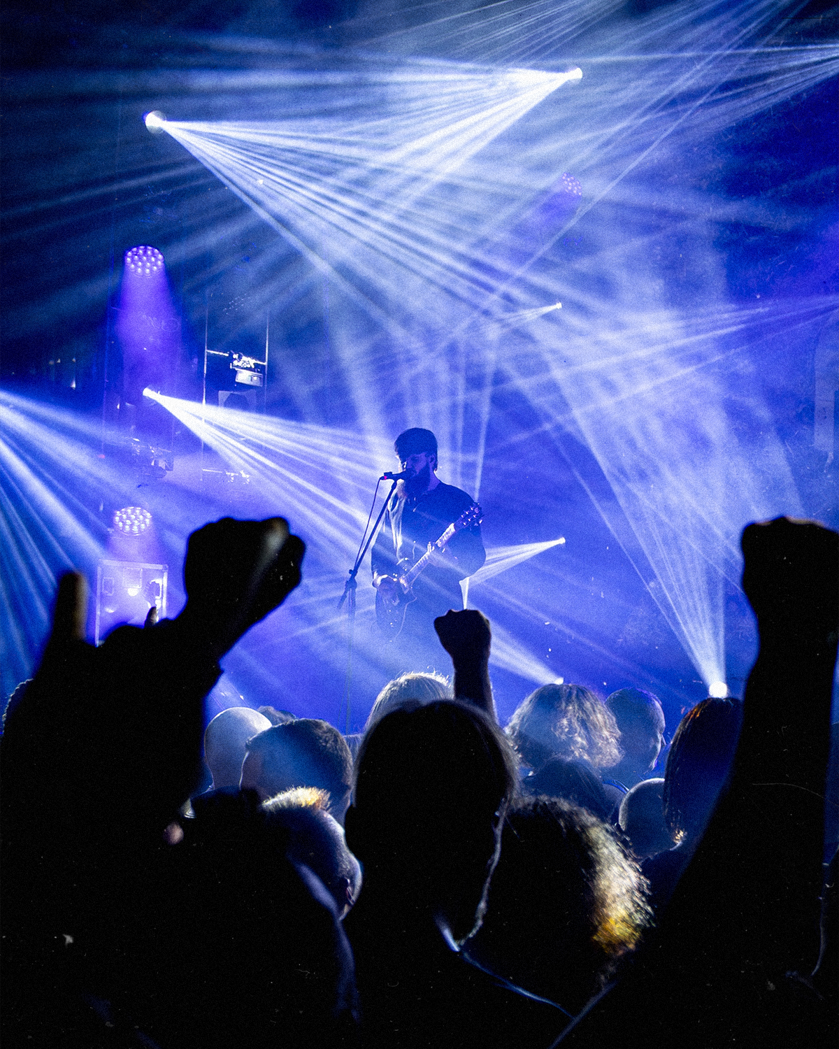 A musician performs on stage with blue and white stage lights shining around him, an audience with raised fists in the foreground.