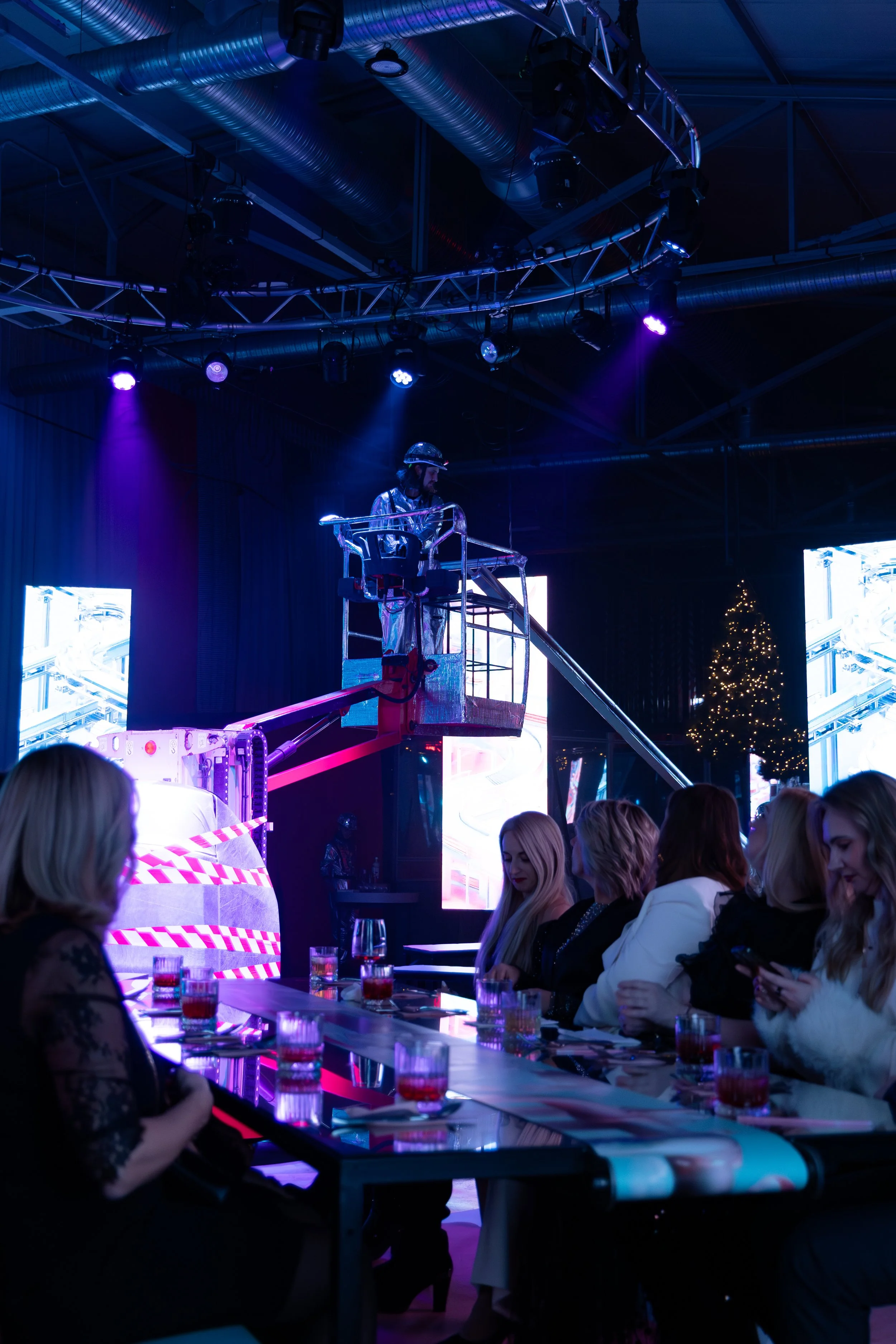 People seated at a long table watching a stage performance, with a man in a metallic suit and helmet on a raised platform, under stage lighting with colorful LED panels and a Christmas tree in the background.