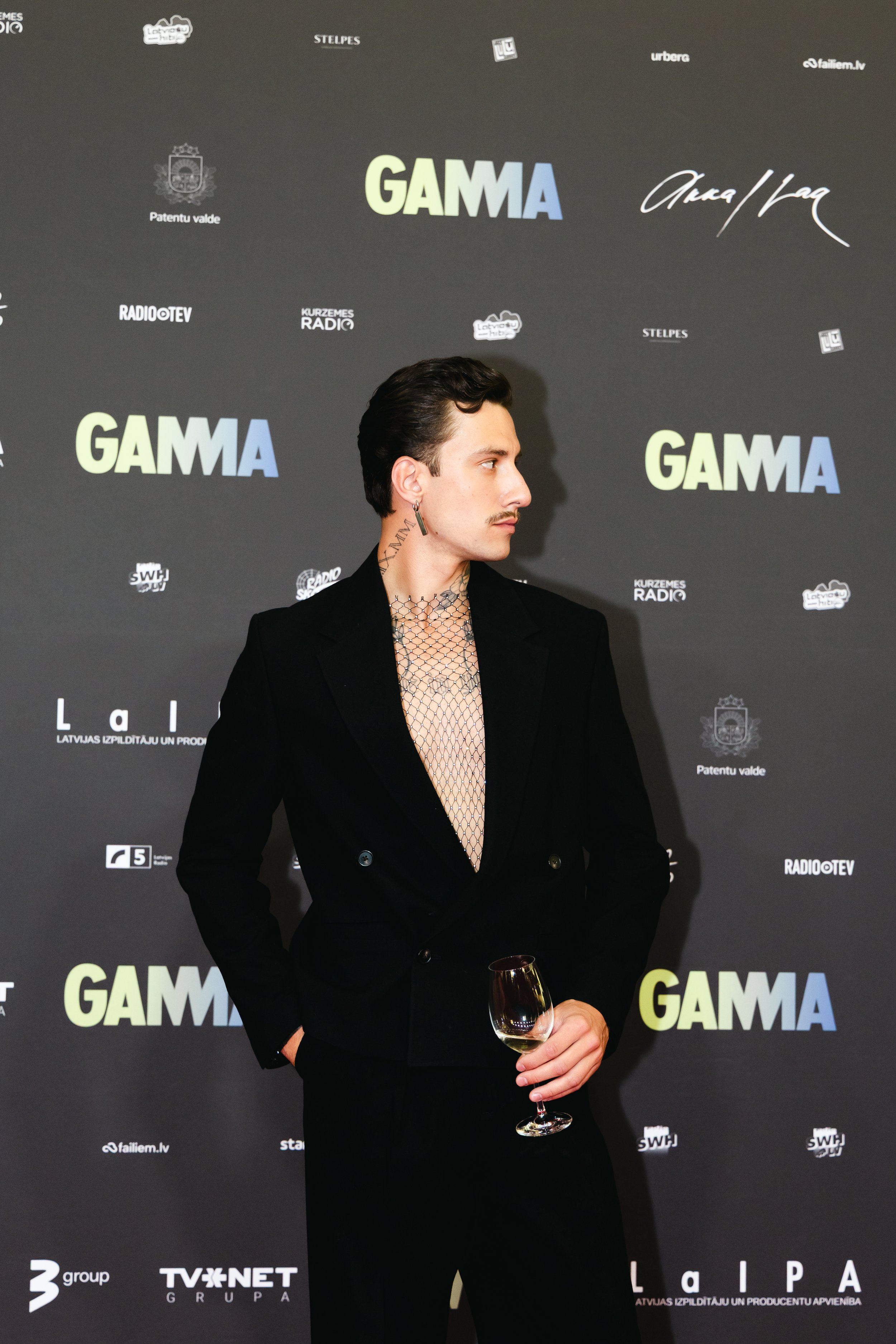 A man with dark hair, mustache, and tattoos on his neck and ear, dressed in a black suit, holding a glass of white wine, standing in front of a black backdrop with multiple logos and the word 'GAMMA' displayed prominently.