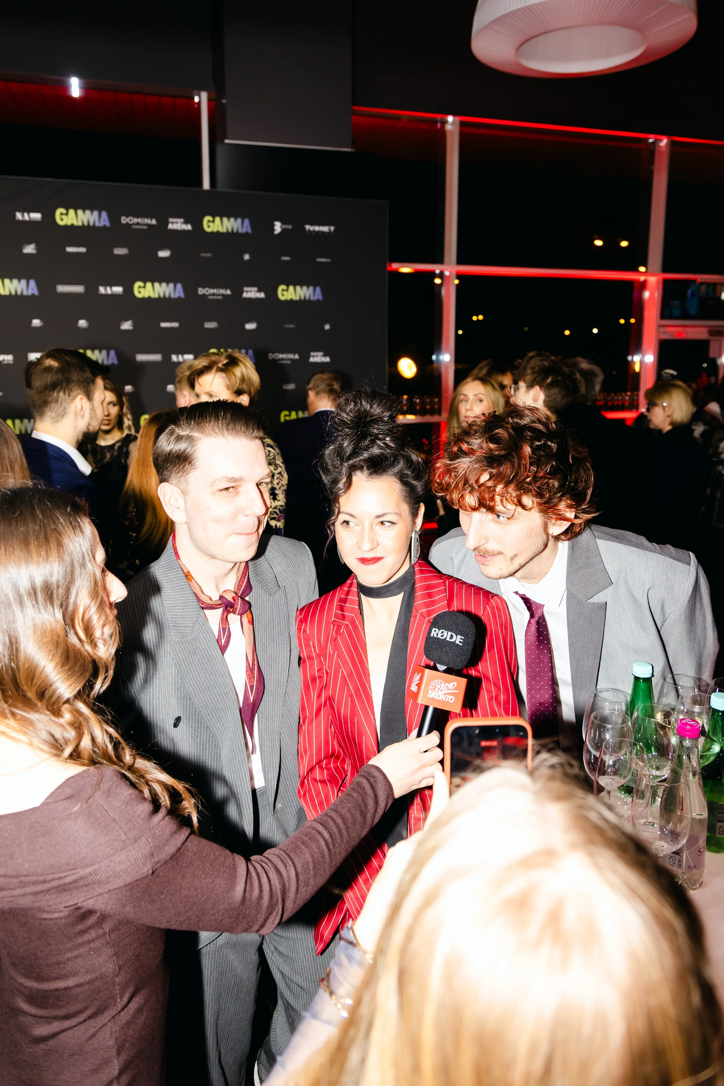 A woman in a red pinstripe suit being interviewed by a podcast host, surrounded by a crowd at an indoor event with a black background, red lighting, and a backdrop with the words "Gamma" and other event sponsors.