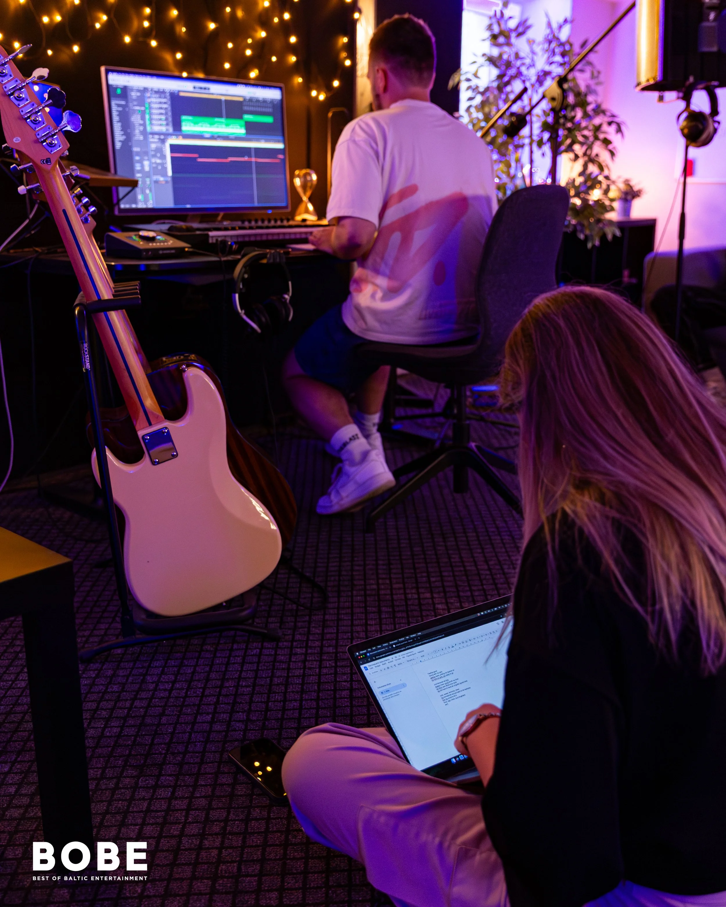 A man wearing a white t-shirt and beige pants sitting on a stool in a music studio, working on a computer with digital audio workstation software on the monitor. A guitar is resting against the desk, and there are headphones hanging from the desk. Th