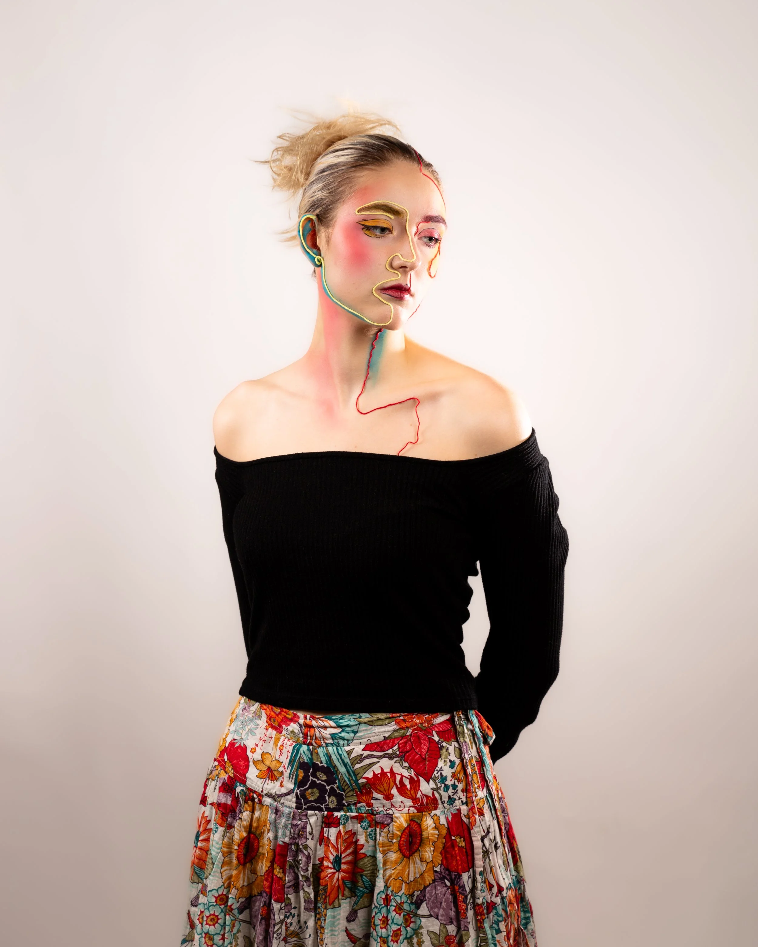 A woman with abstract face and body paint, wearing a black off-the-shoulder top and a floral skirt, standing against a plain light background.