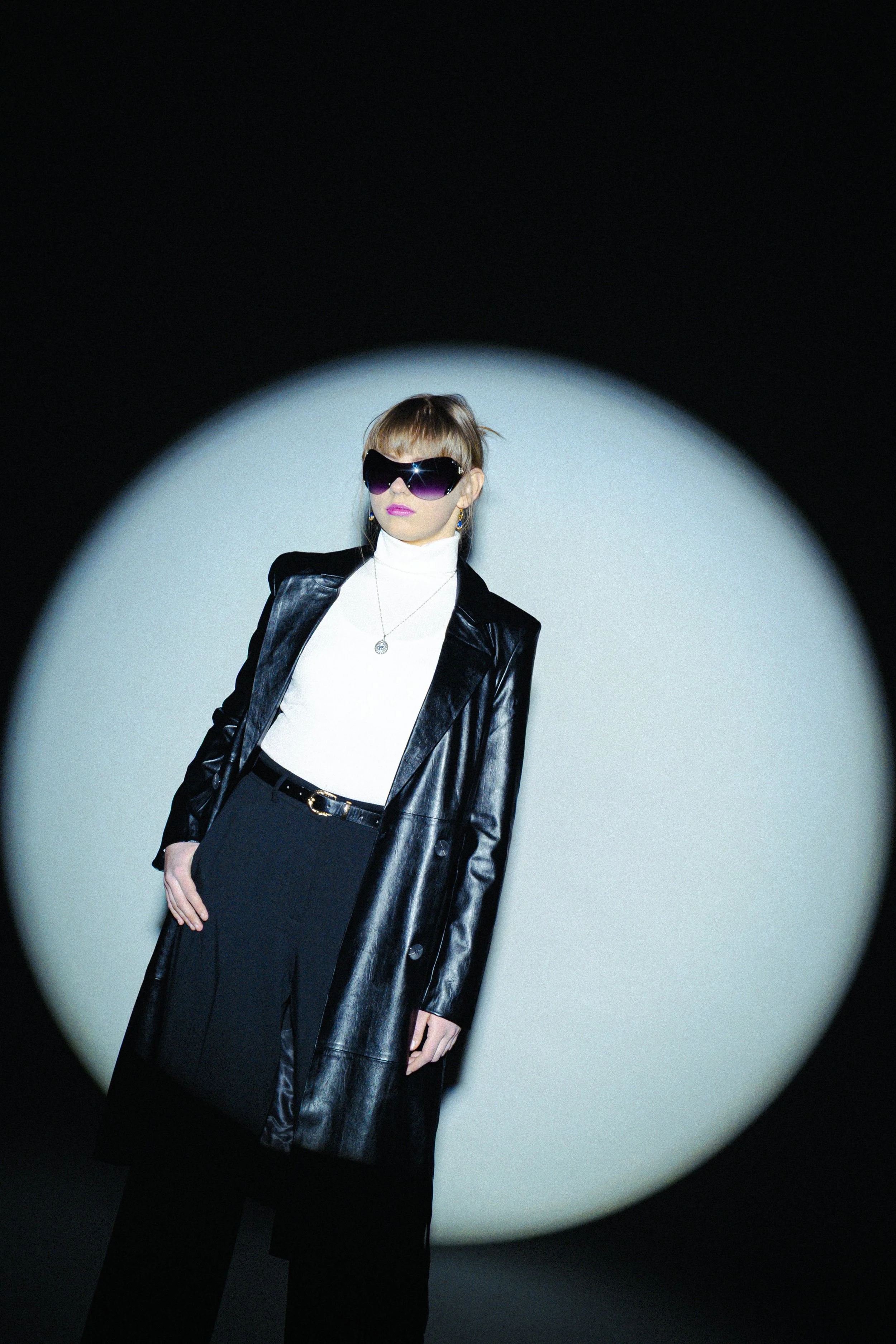 A woman standing in front of a spotlight, wearing oversized sunglasses, a white turtleneck, a black leather jacket, and black pants, in a dark setting
