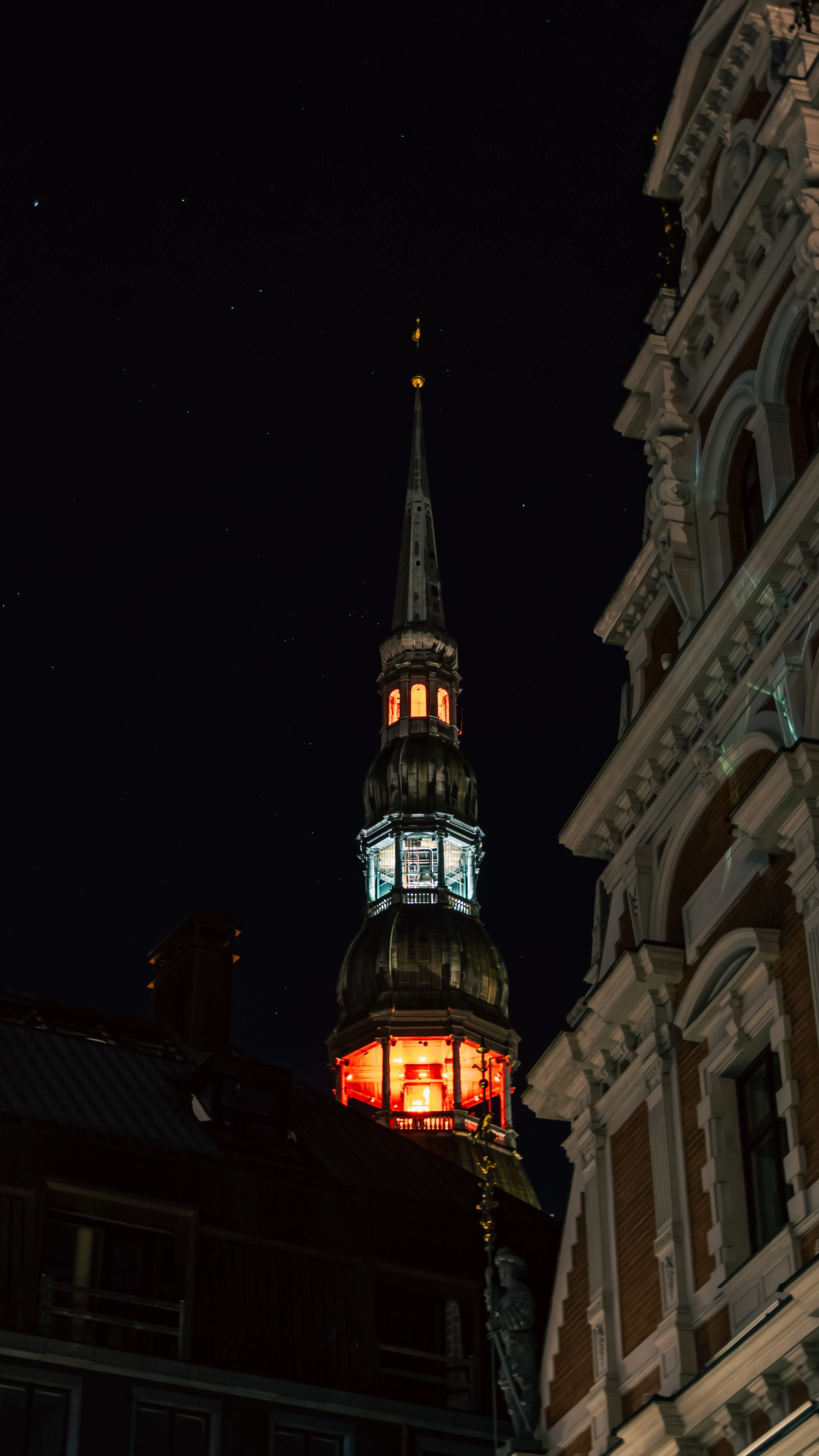 Nighttime view of a tall, ornate church steeple illuminated with red lights, against a starry sky, with historic buildings nearby.