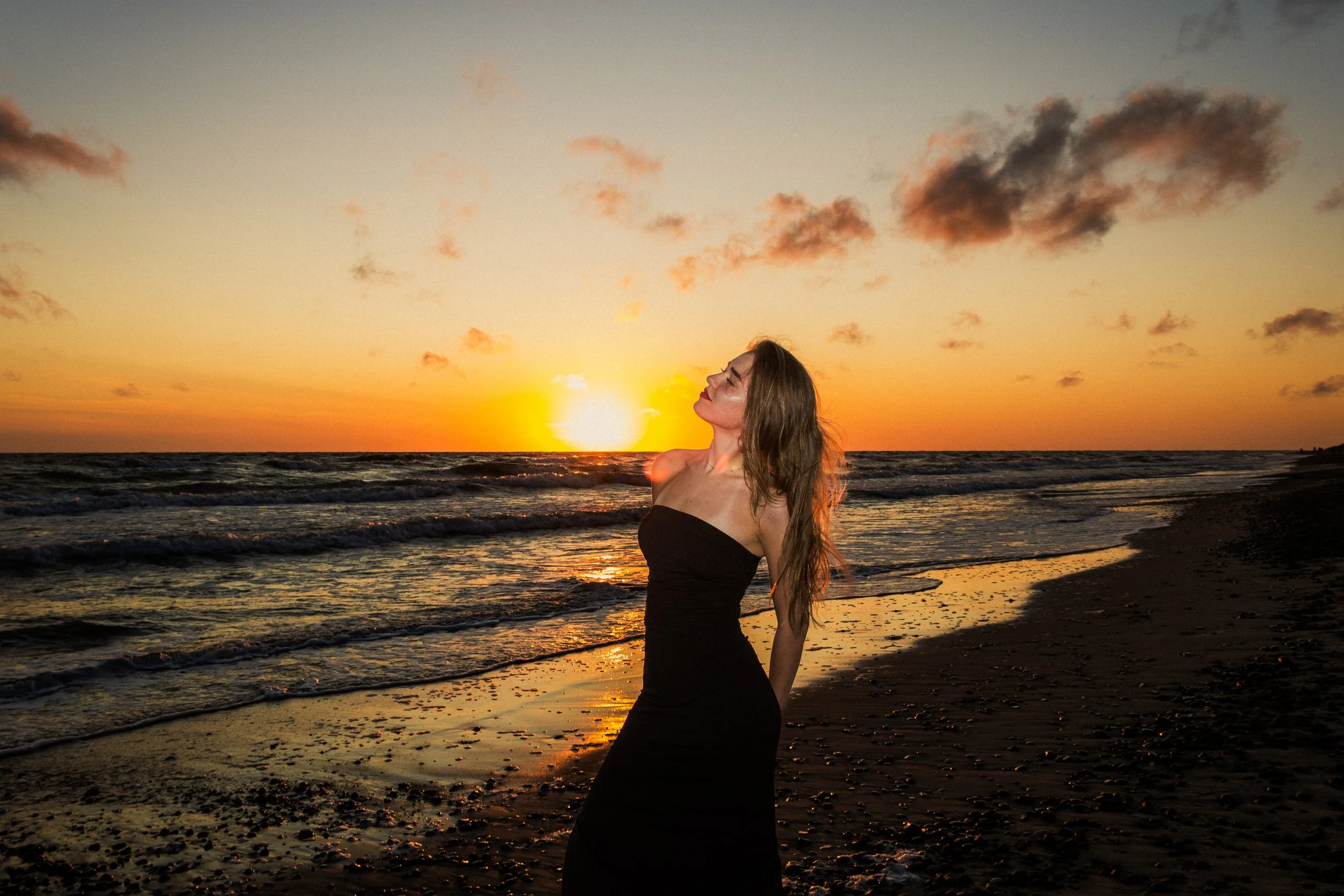 A woman with long brown hair standing at the beach during sunset, wearing a black strapless dress, with the ocean and sky with clouds in the background.