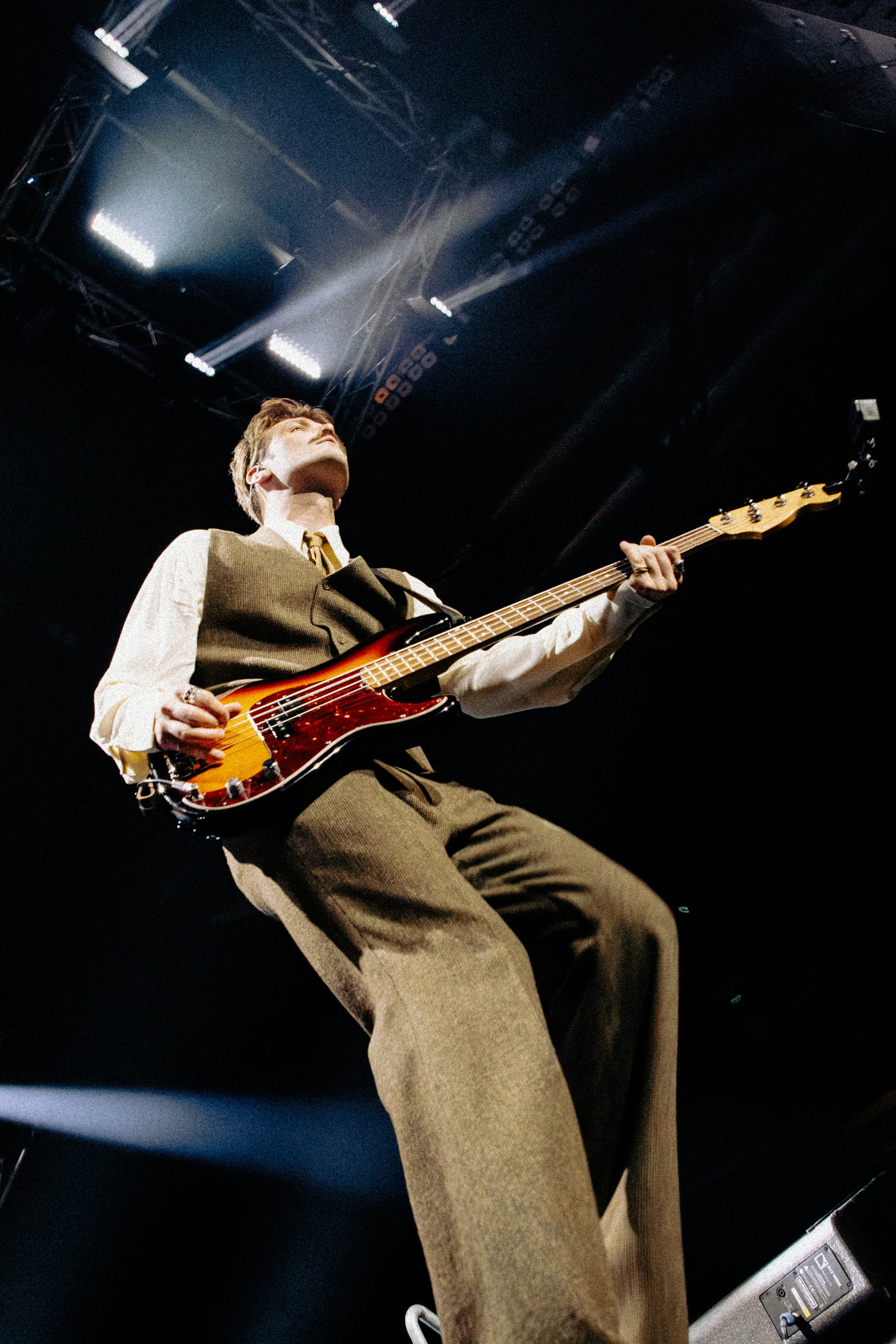 A person wearing a beige vest and dress shirt playing an electric bass guitar on stage with stage lights shining from above.