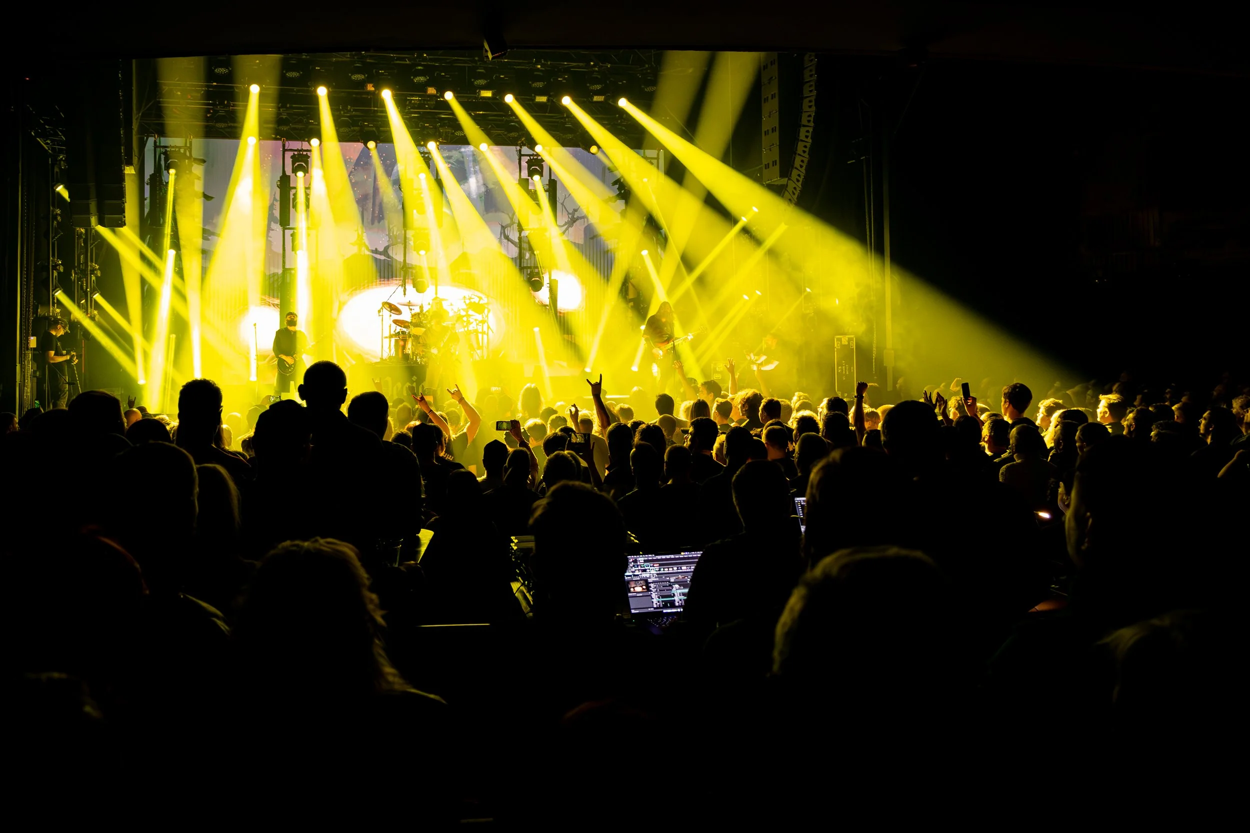 Concert audience viewing a performance on stage with yellow lighting, with a sound technician working at a mixing console in the foreground.