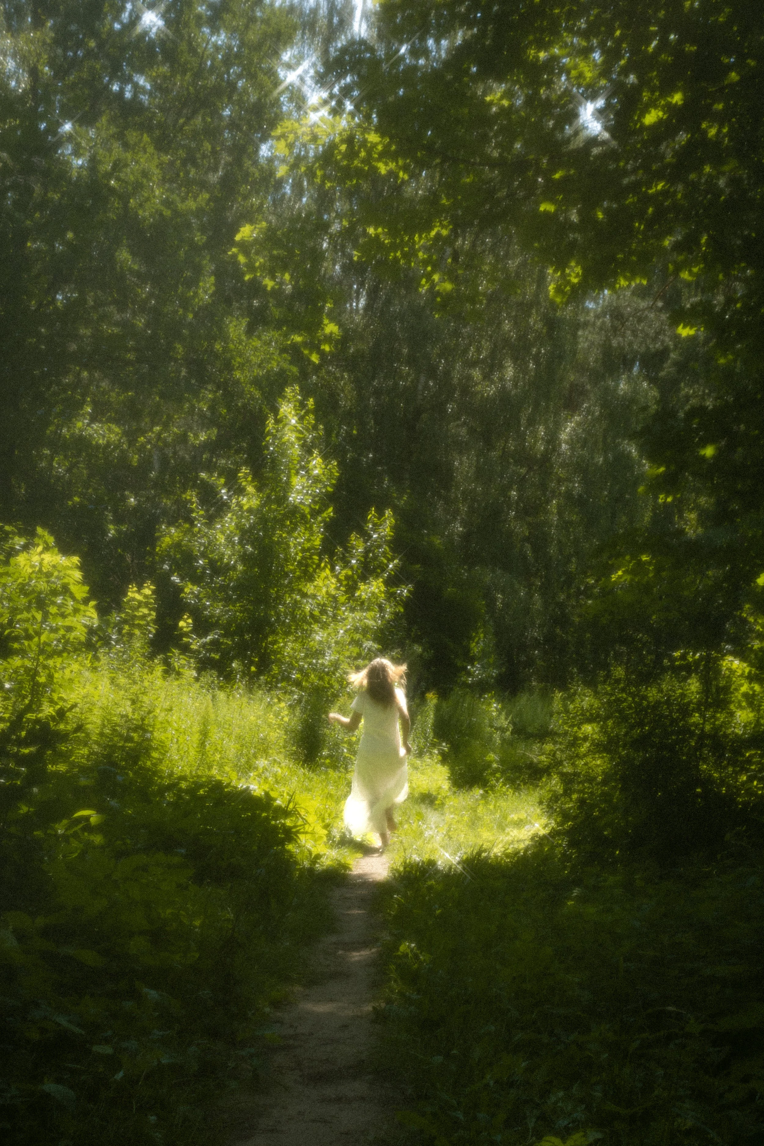 A woman in a white dress walking along a forest trail, surrounded by lush green trees and sunlight filtering through the leaves.