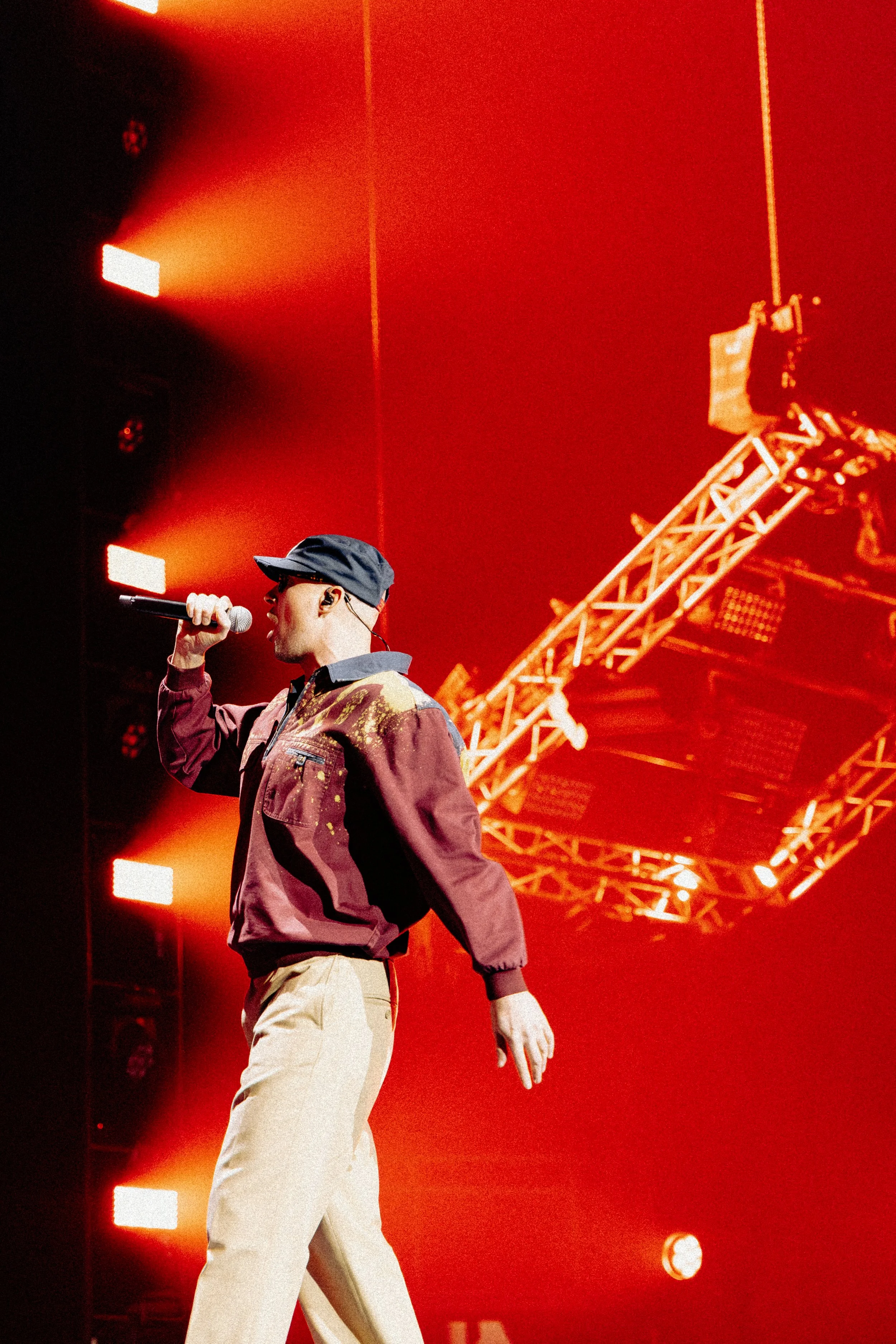 A person speaking into a microphone on stage with bright orange and red lighting, wearing a dark blue cap, glasses, a maroon jacket, and beige pants, with stage equipment and lighting structures in the background.