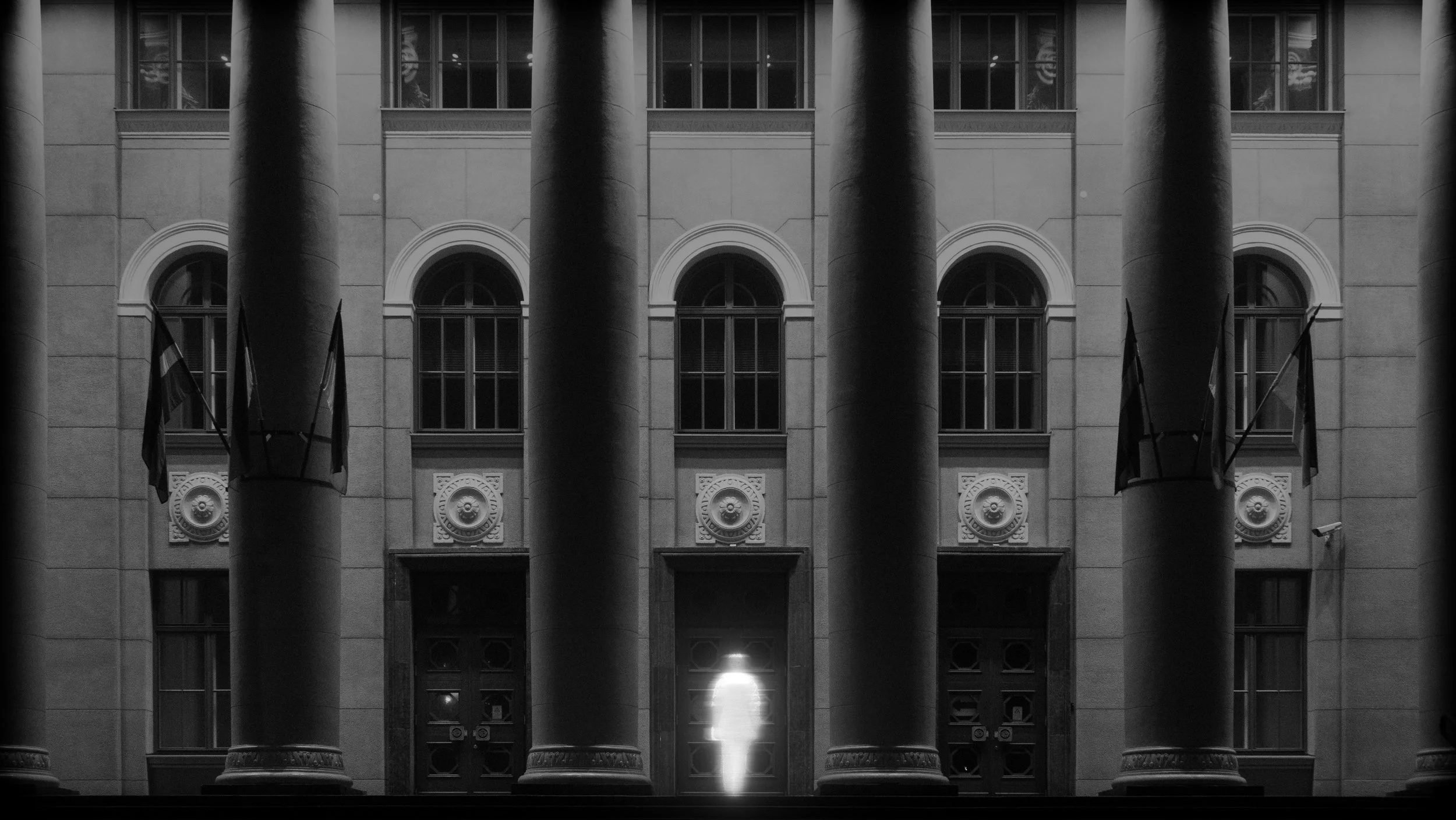 Night view of a building with large columns and flags, illuminated by a light at the bottom center.
