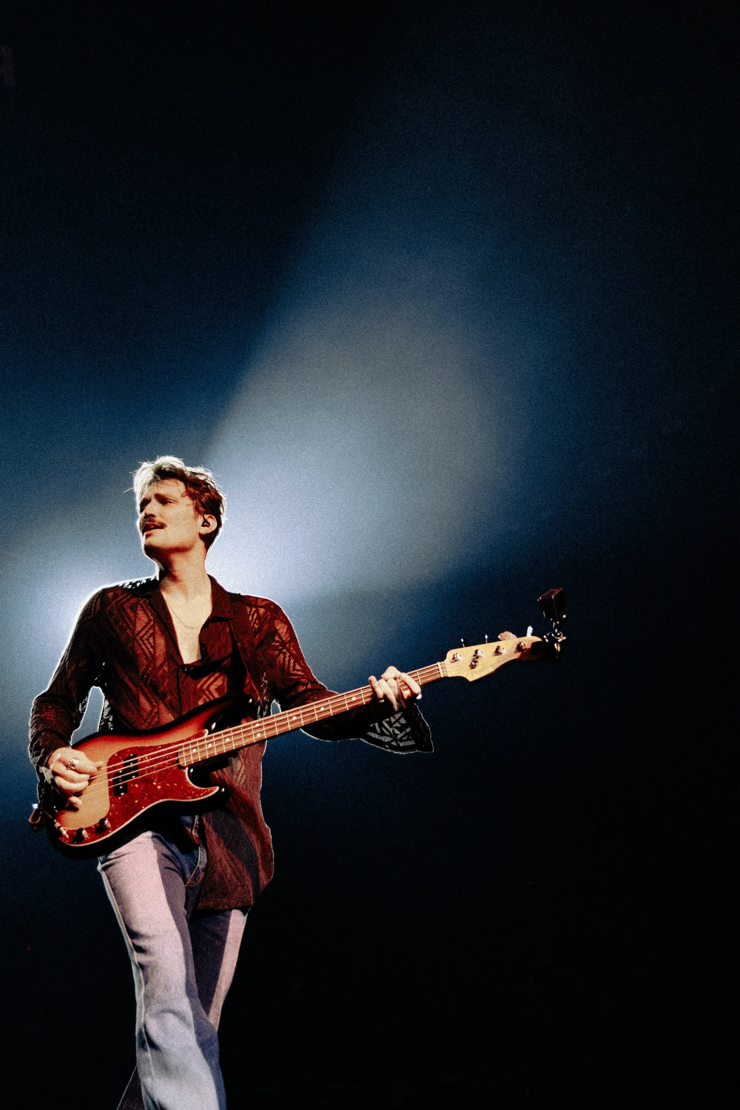 A man playing an electric guitar on stage, wearing a patterned shirt and light-colored pants, with dark background and stage lighting.