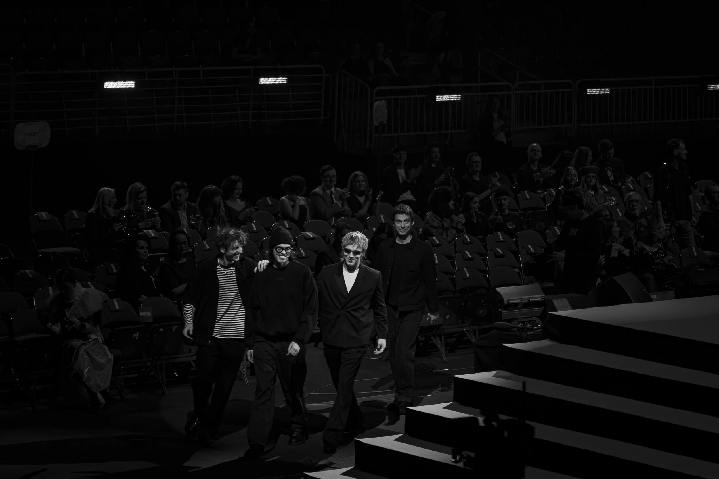 Group of people walking across a stage at an event, with some audience members seated in the background.