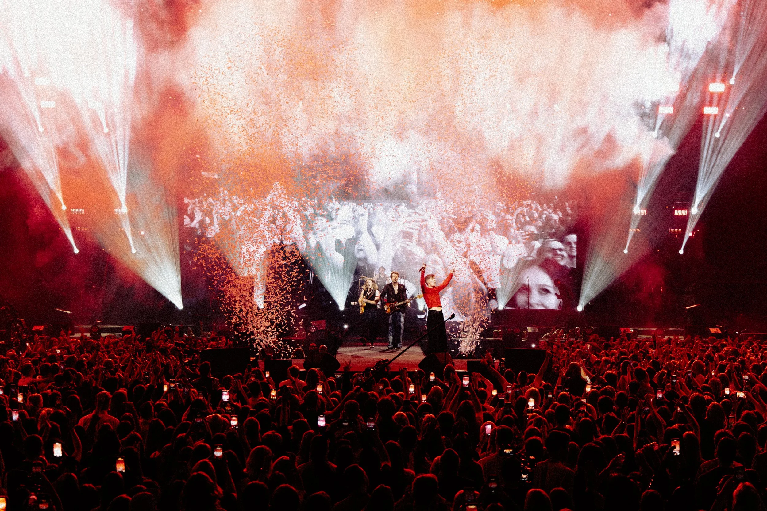 Concert stage with musicians performing, large crowd with many people holding smartphones, bright stage lights, and confetti in the air.