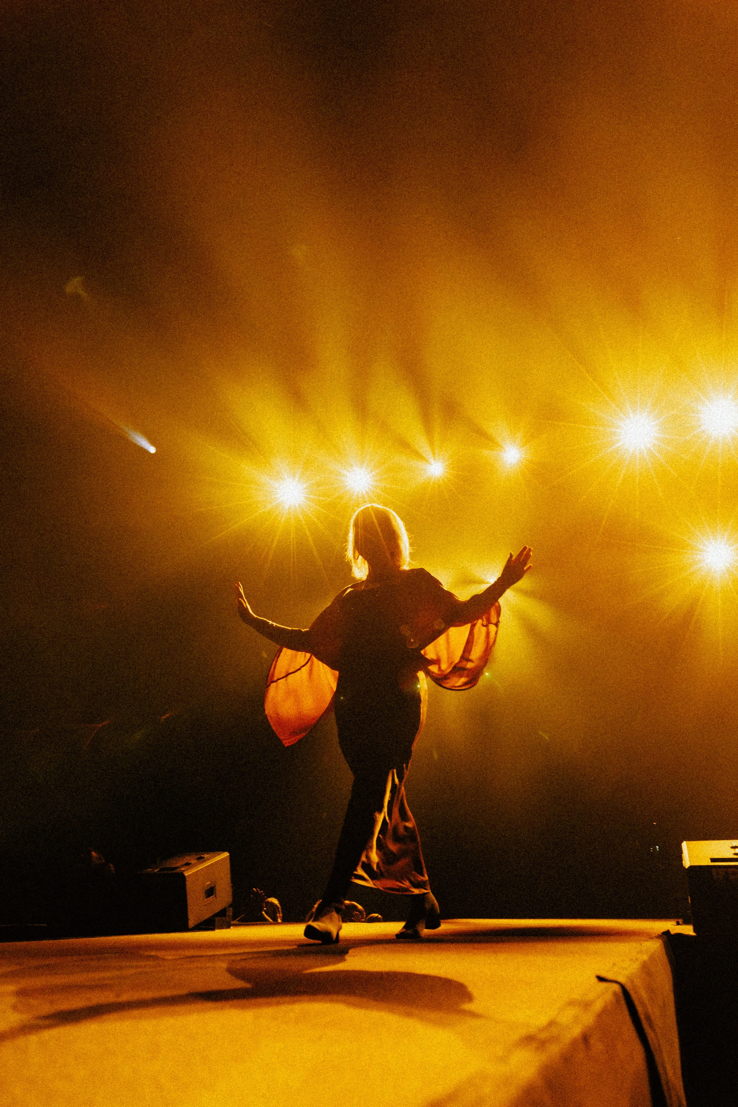 A performer on stage dancing with arms raised, illuminated by bright stage lights in warm colors, with an audience and equipment visible in the foreground.