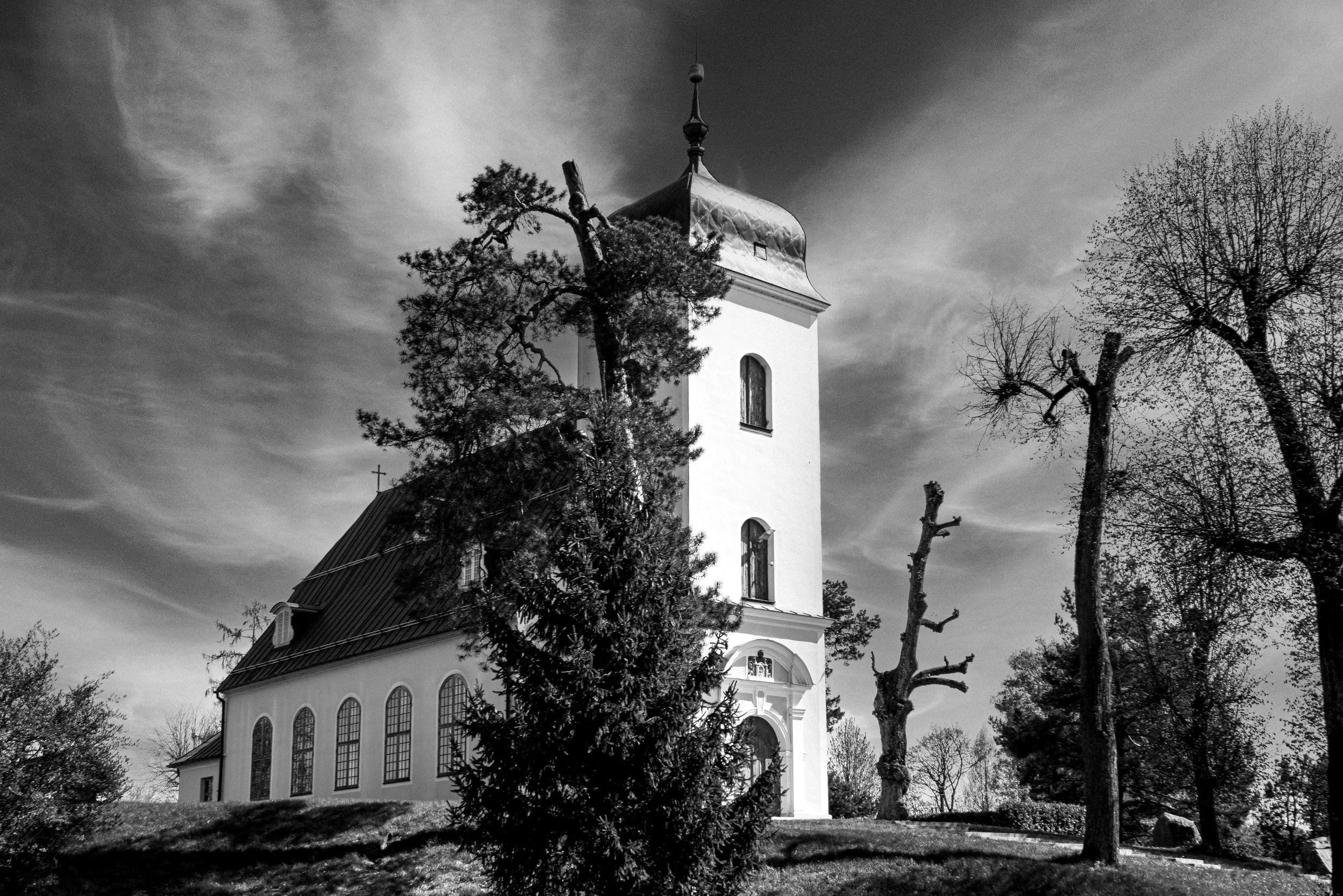 A white church with tall, arched windows and a steeple, partially obscured by detailed, leafless trees in a black-and-white photo.