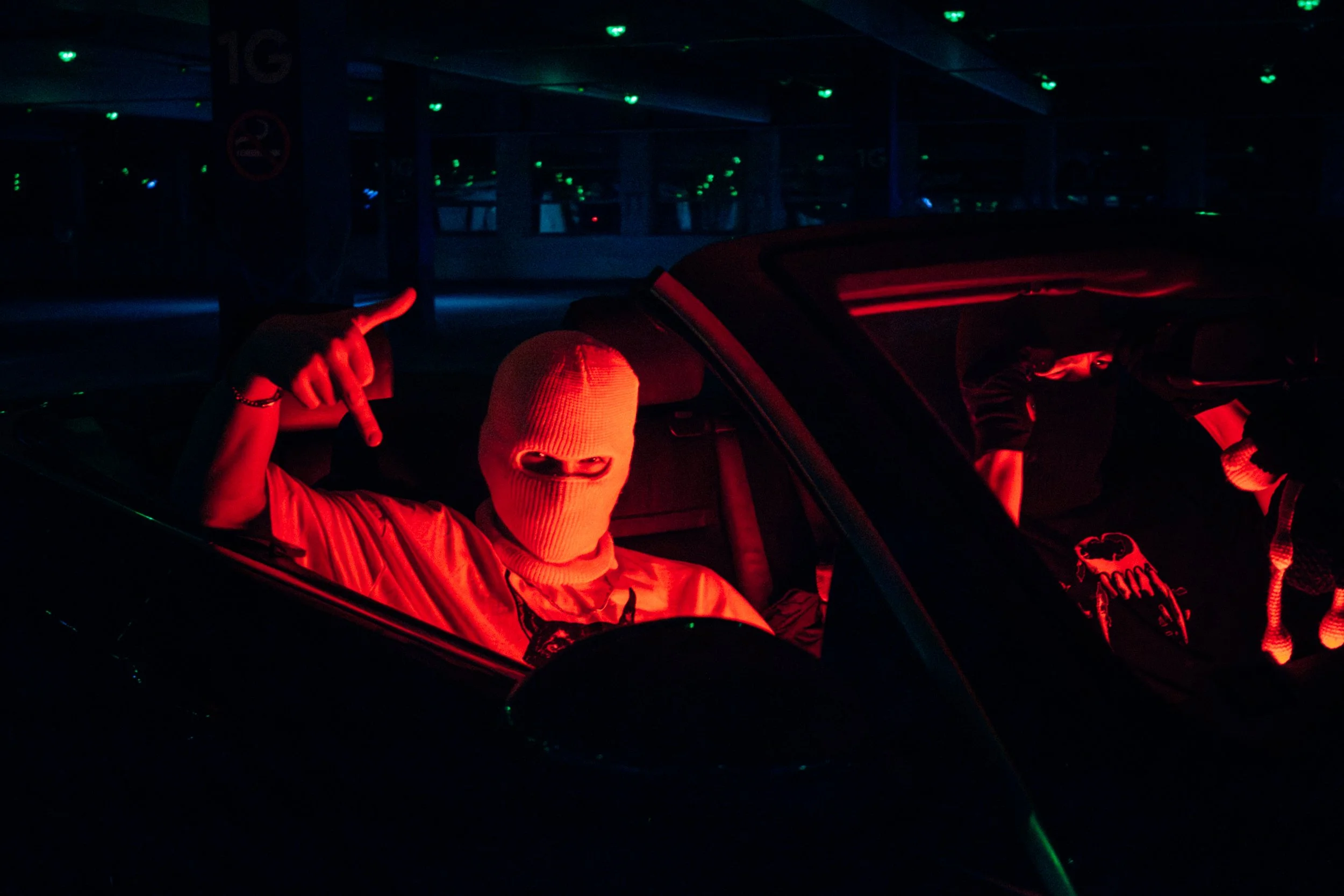 Two individuals in a dark parking garage, one wearing a ski mask and orange jacket, pointing with their hand, with green and red lights reflecting off the scene.